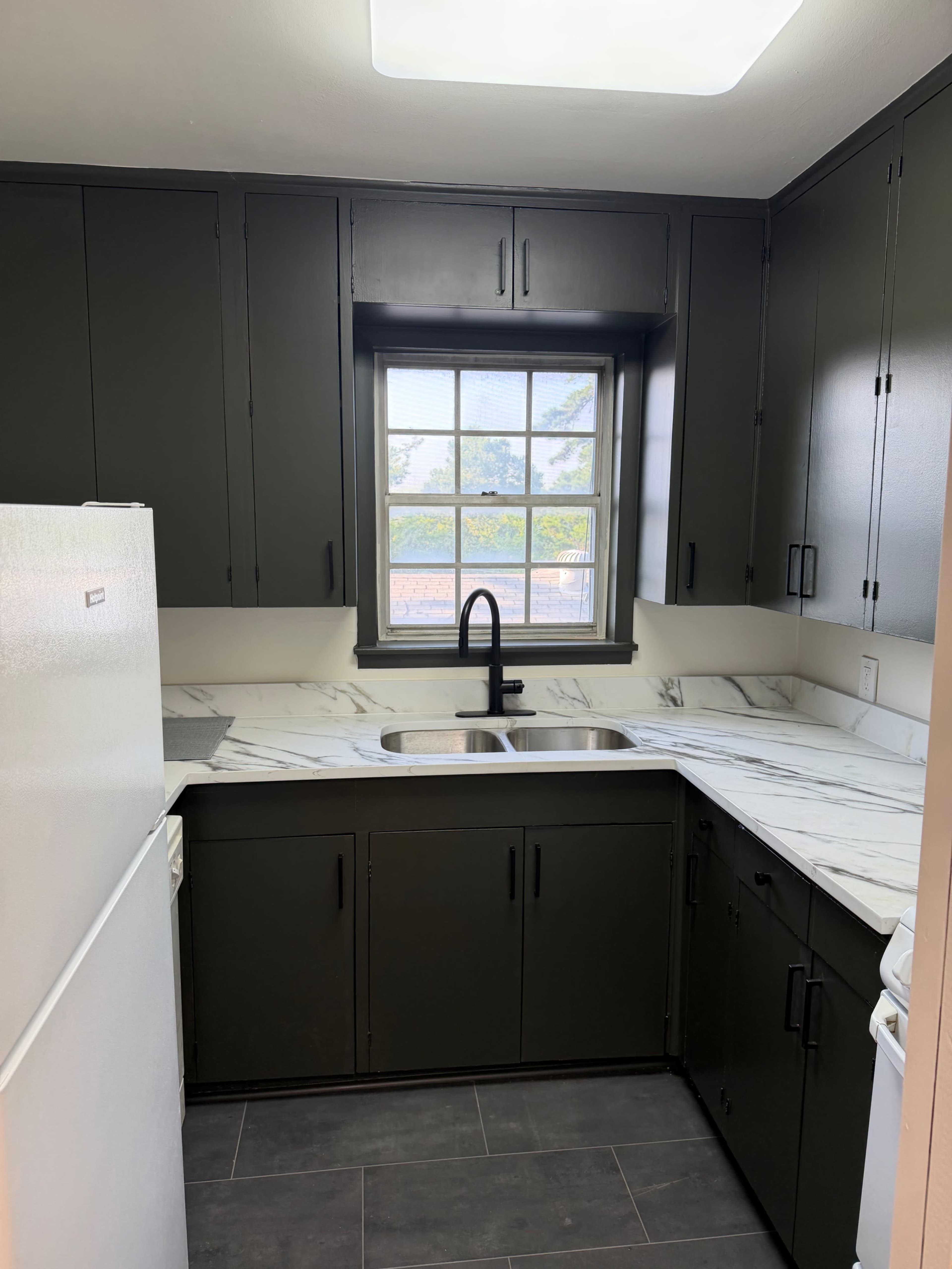 The image shows a modern kitchen with dark cabinetry, a marble countertop, and a black faucet installed over a double sink, illuminated by natural light through a window.