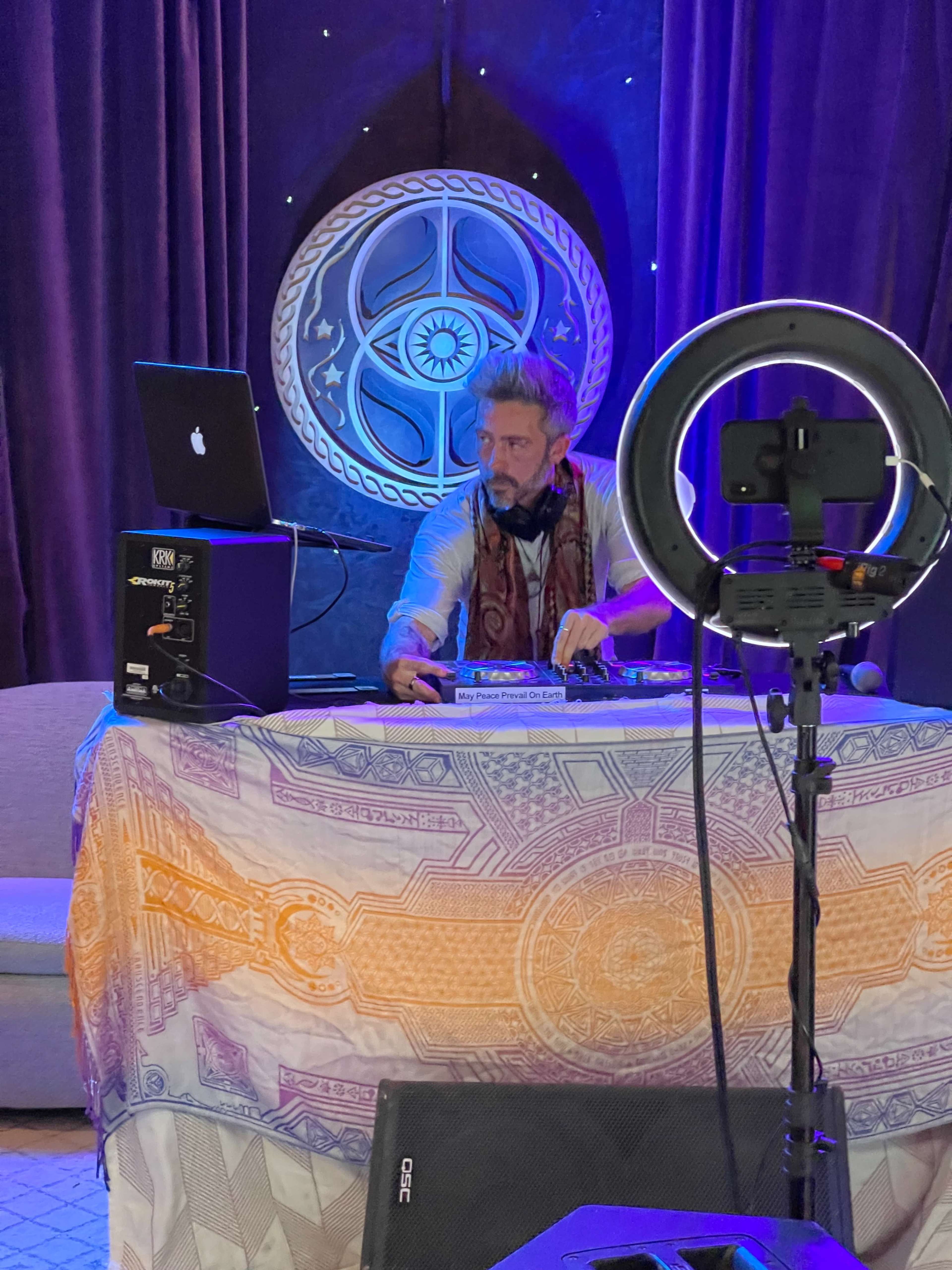 A DJ operates a laptop and turntables behind a decorated table with a backdrop featuring a circular design and purple curtains.