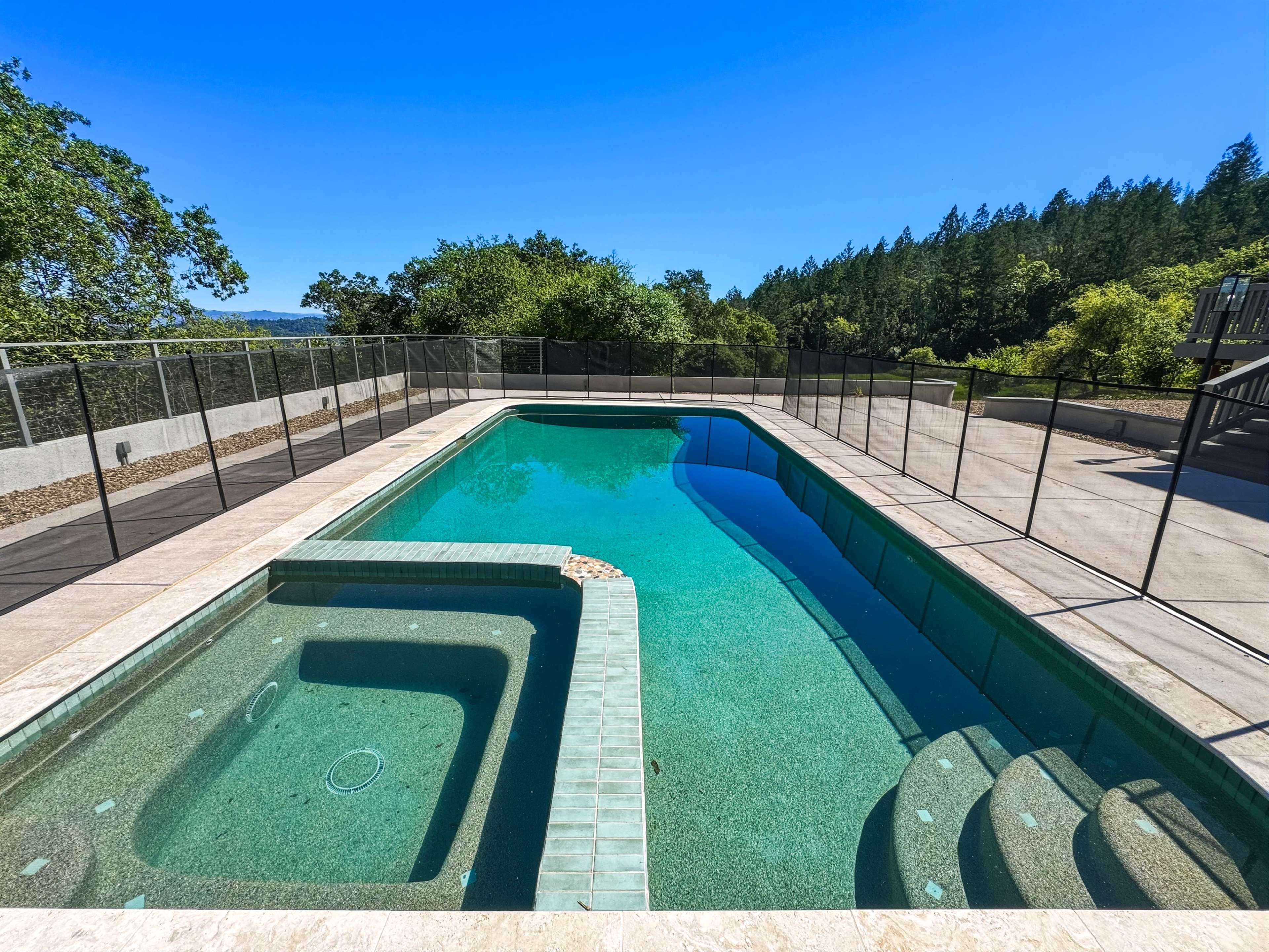 The image shows a rectangular swimming pool with a spa section, surrounded by a safety fence and overlooking a wooded area under a clear blue sky.