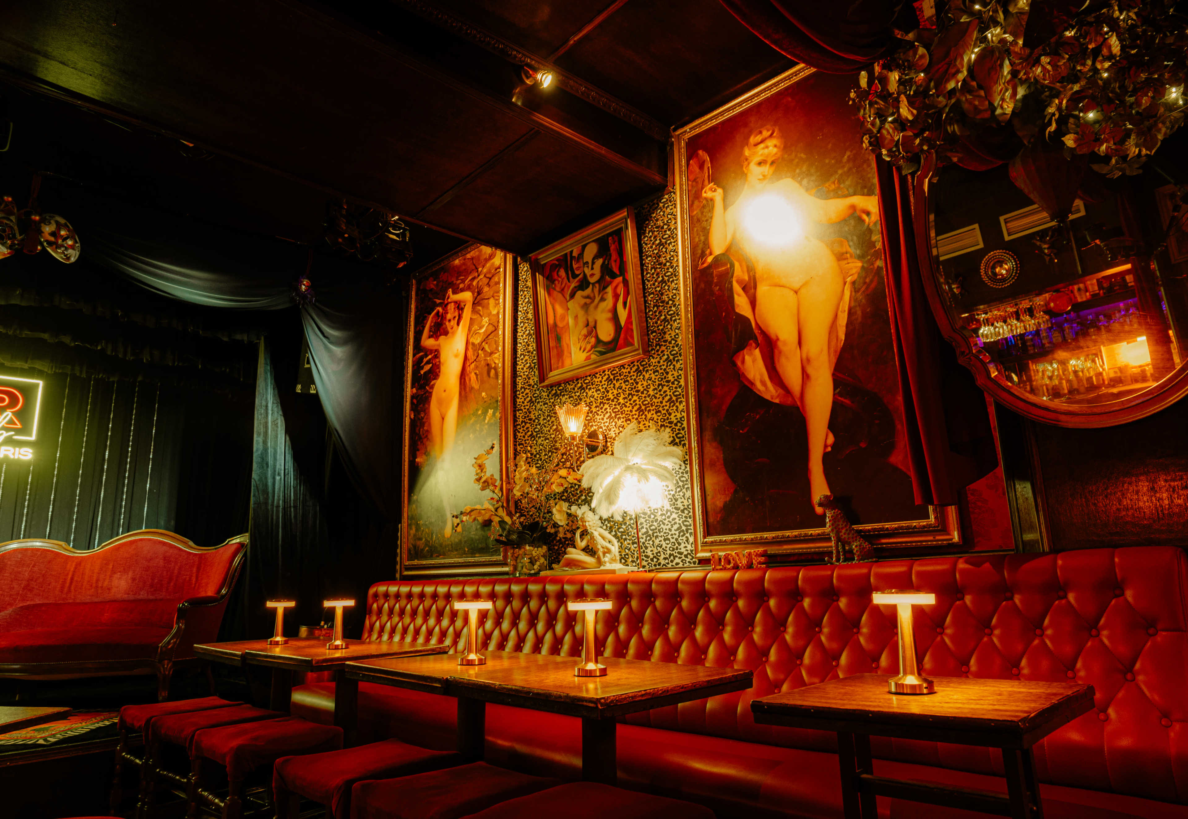 The image shows a dimly lit lounge with red velvet seating, decorative wall art, and tables illuminated by small lamps.