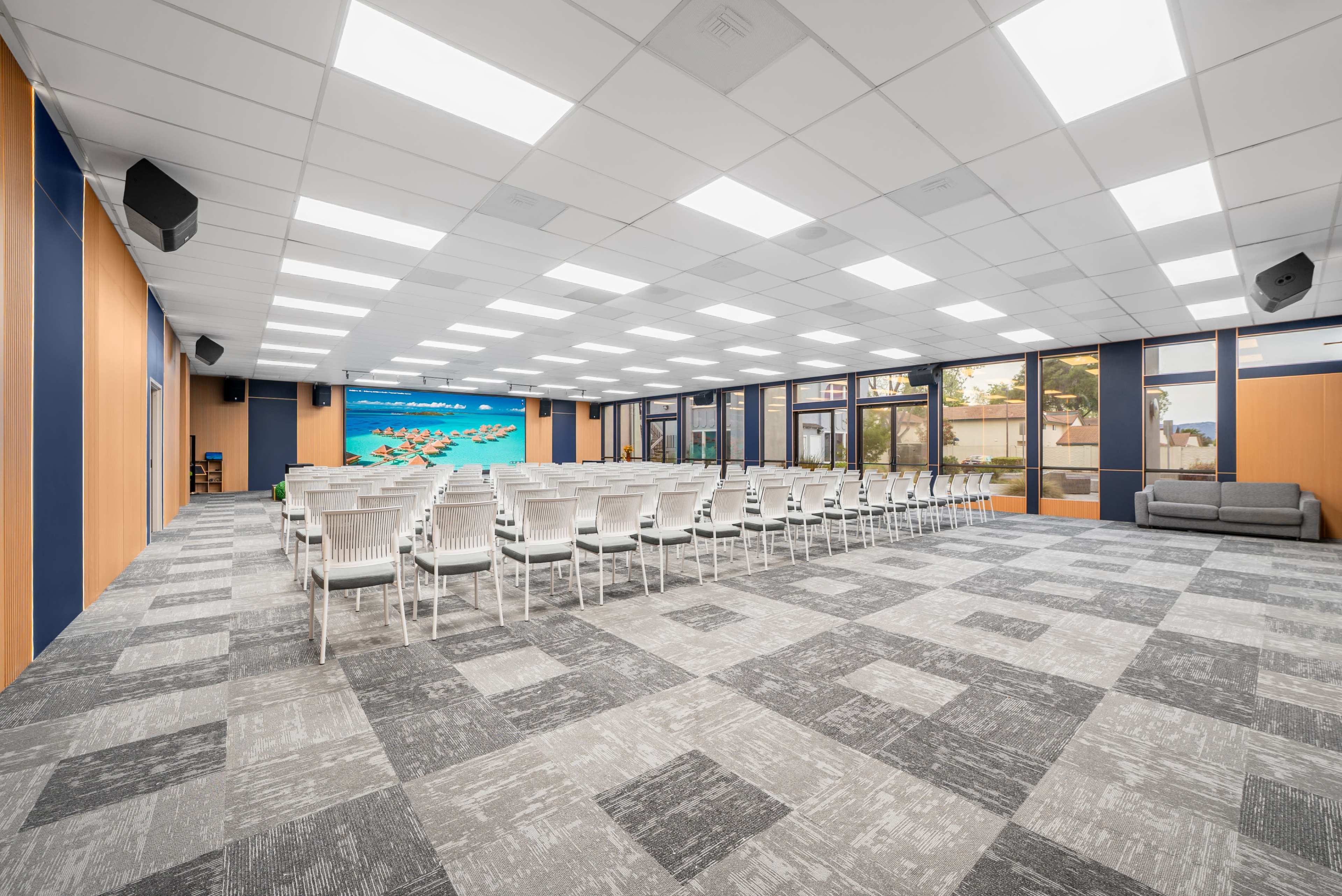 The image shows a spacious conference room arranged with rows of white chairs facing a large digital screen, with large windows allowing natural light to enter.