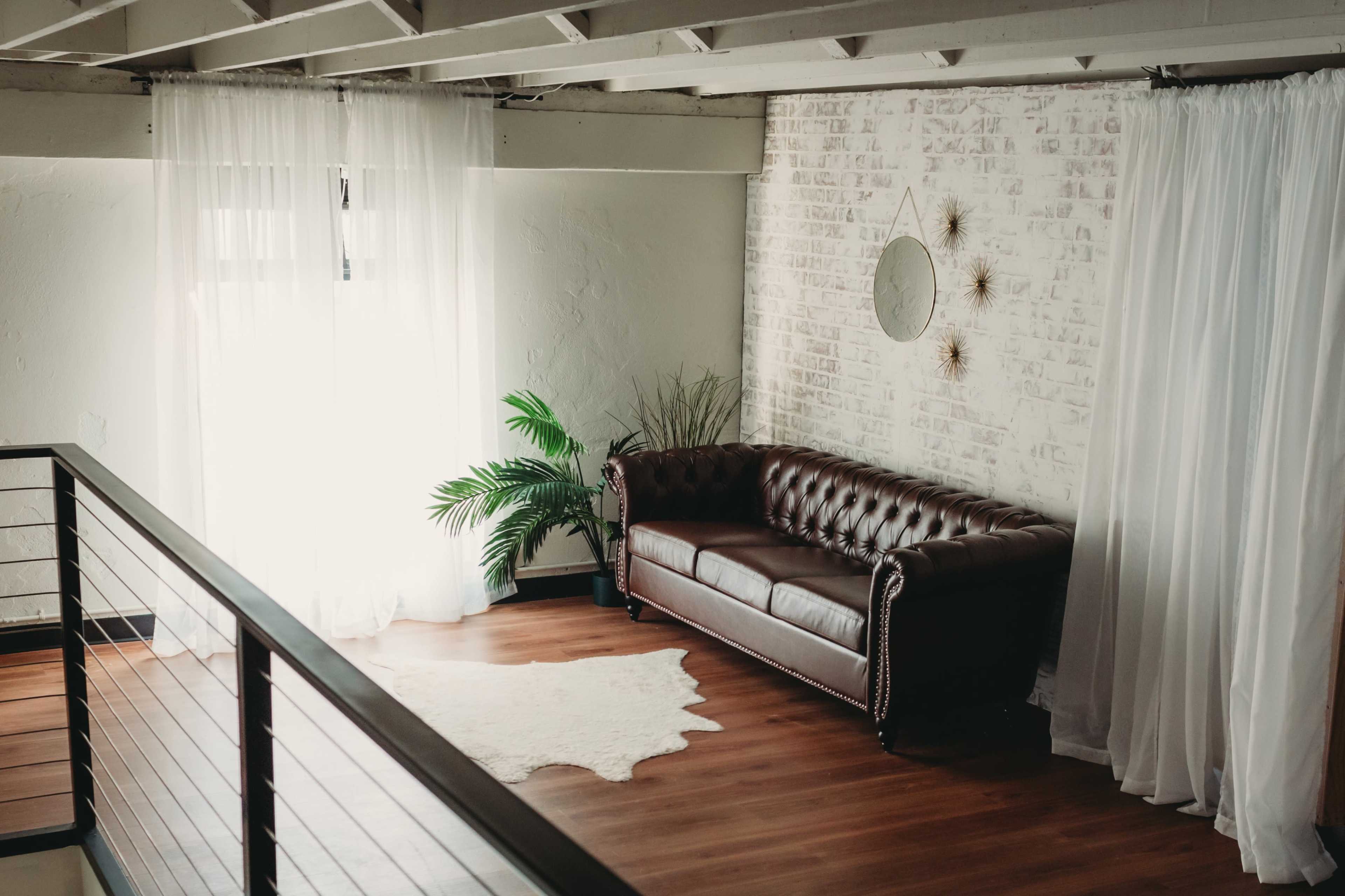 A spacious loft with a brown leather sofa, a large window draped with sheer curtains, and a decorative wall mirror.