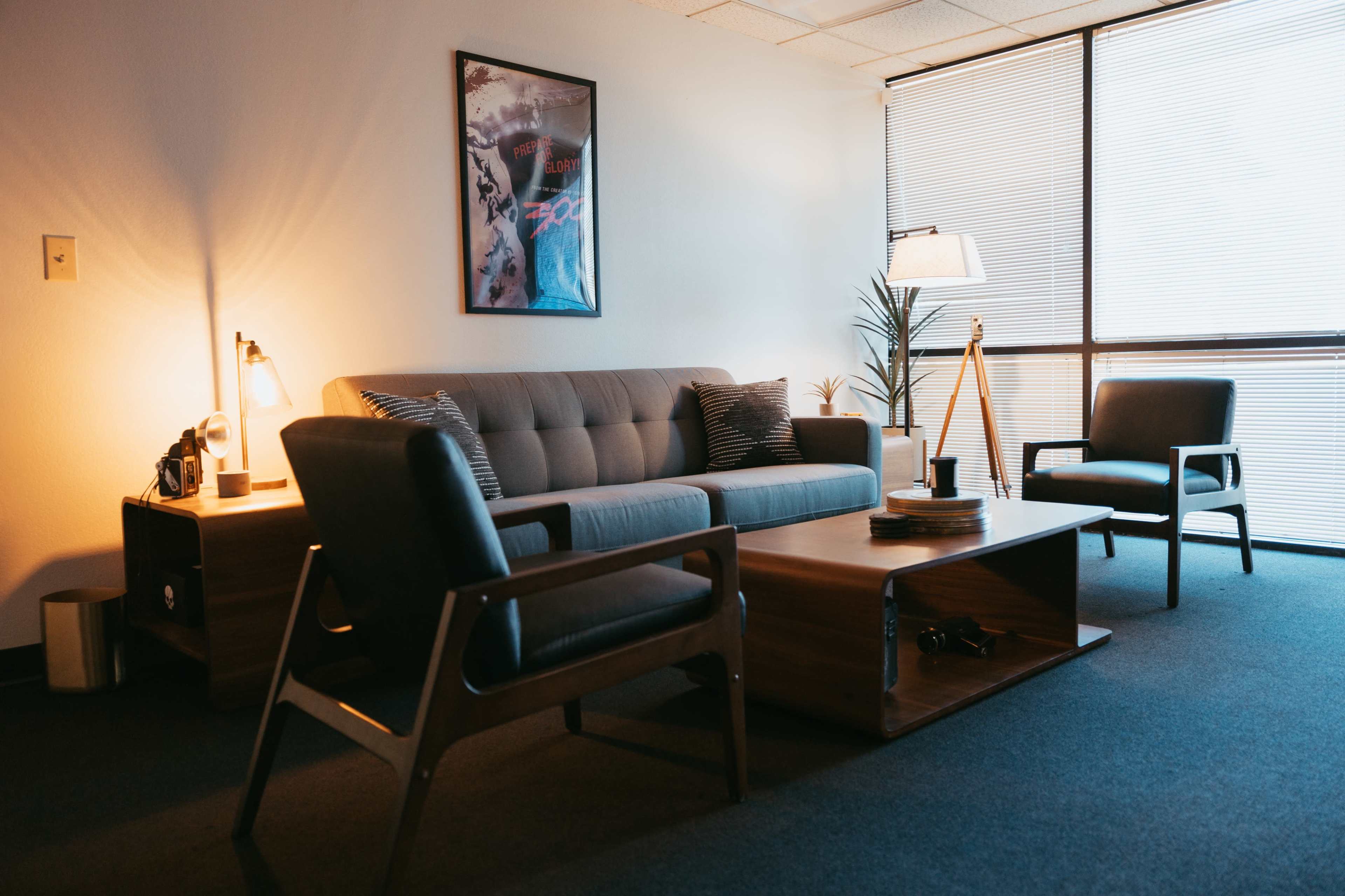 A modern office seating area features a gray couch, two black armchairs, a wooden coffee table, and a lamp, illuminated by natural light from large windows.