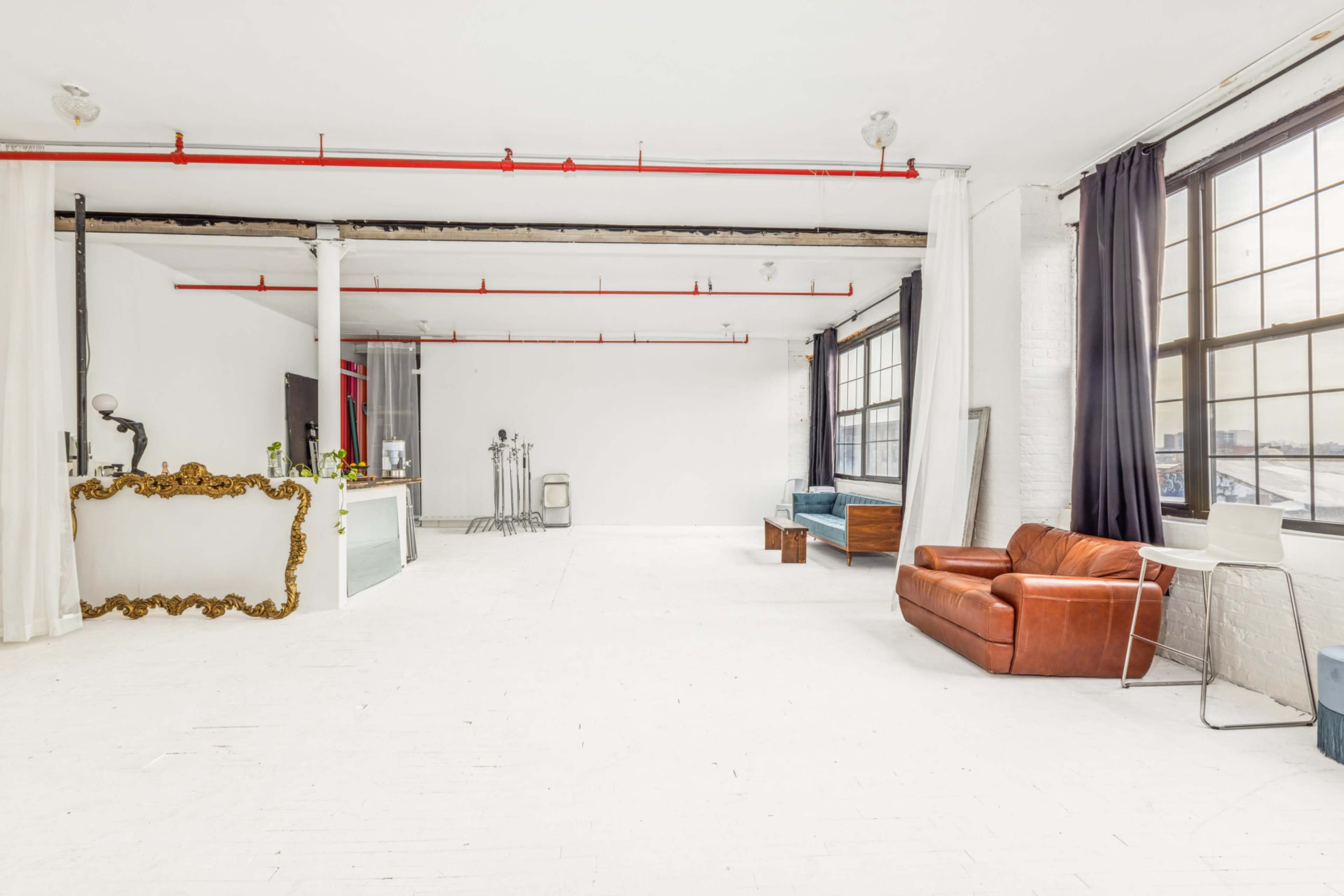 The image shows a spacious, minimalist loft with white walls, large windows, and a mix of furniture, including a brown leather sofa.