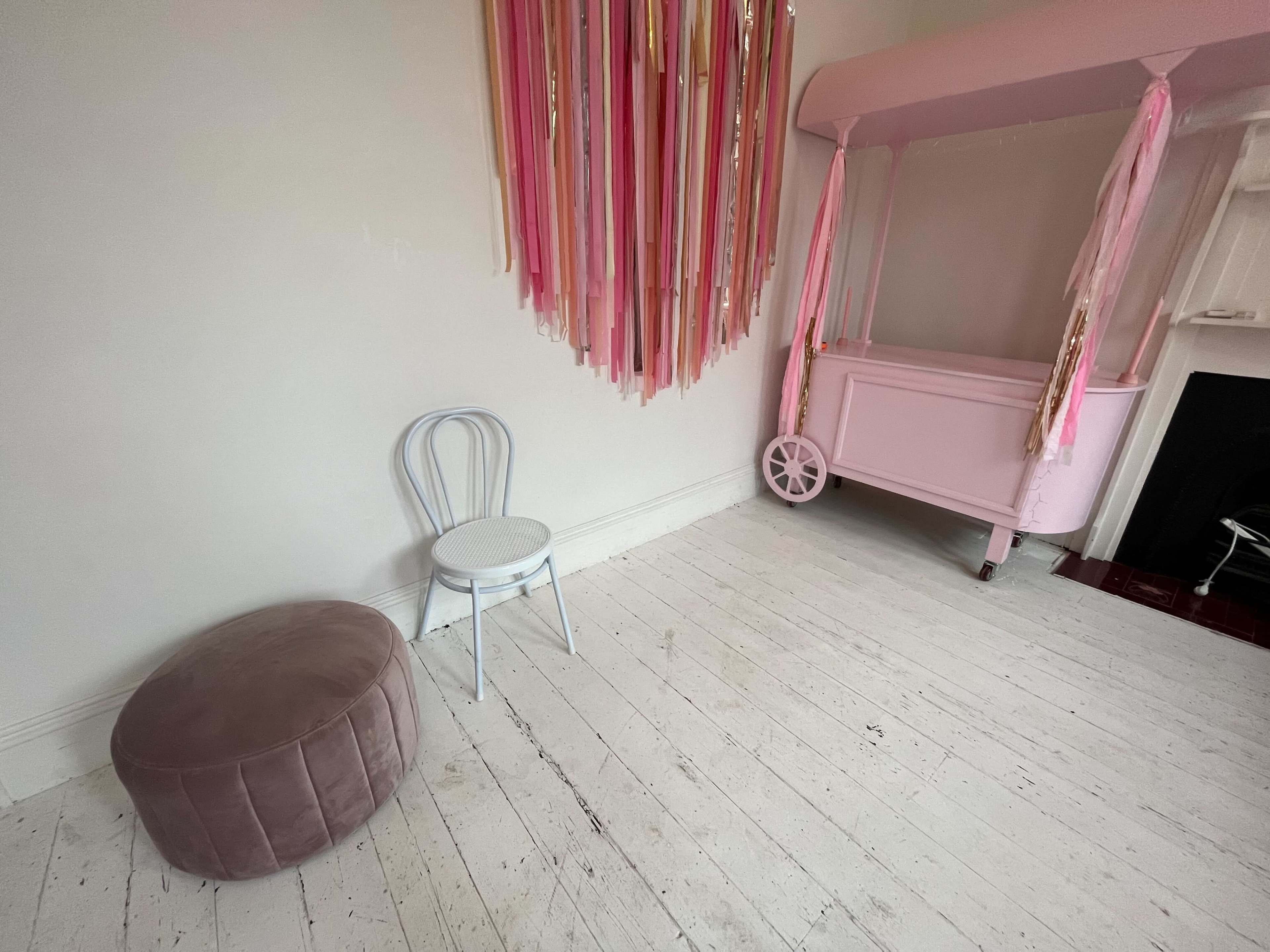 A pale room with a pink cart, a gray chair, and a round pink ottoman on a white wooden floor.