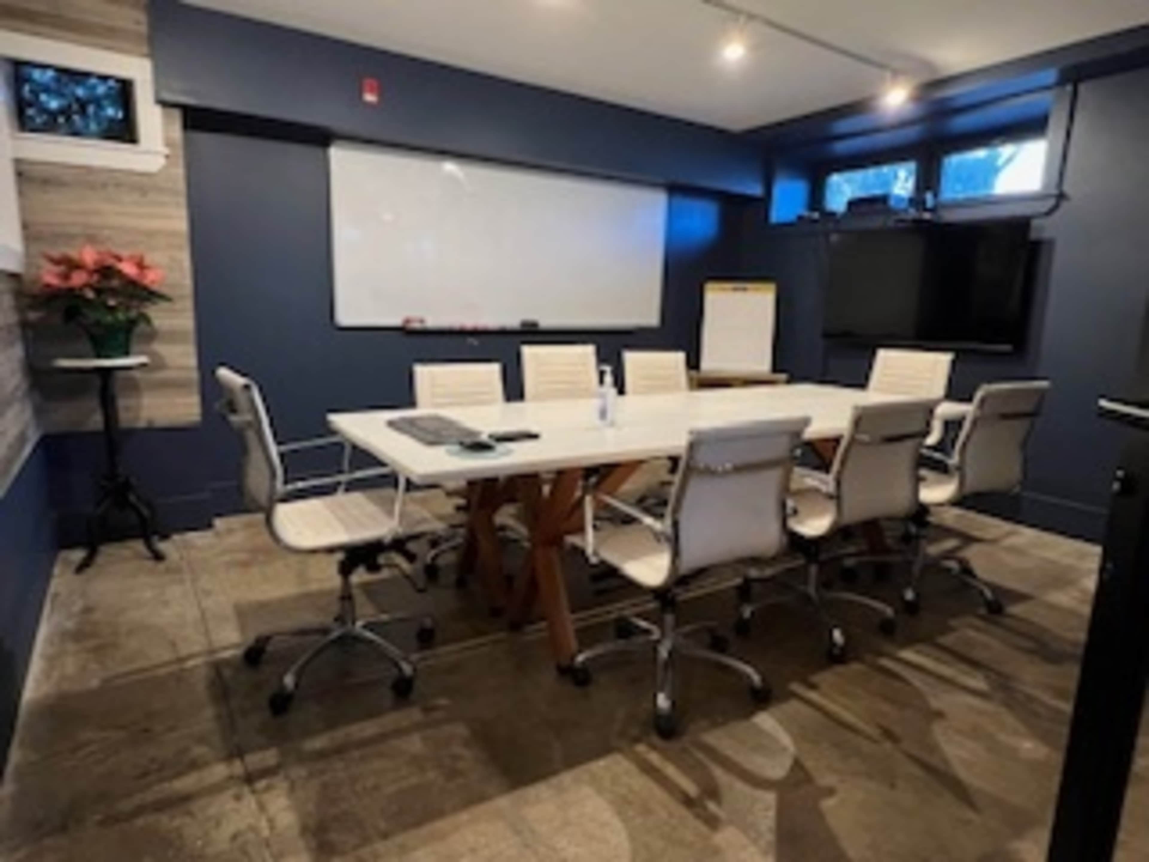 A modern conference room features a large white table surrounded by ergonomic chairs, a whiteboard on the wall, and a TV screen.