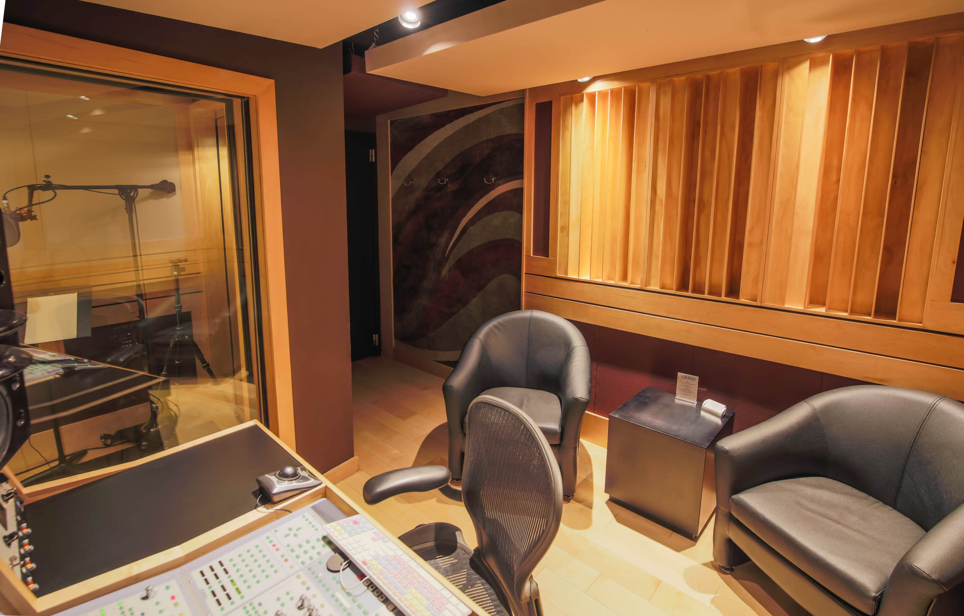 A modern sound recording studio featuring a control desk, two chairs, and a soundproofed vocal booth visible through a glass partition.