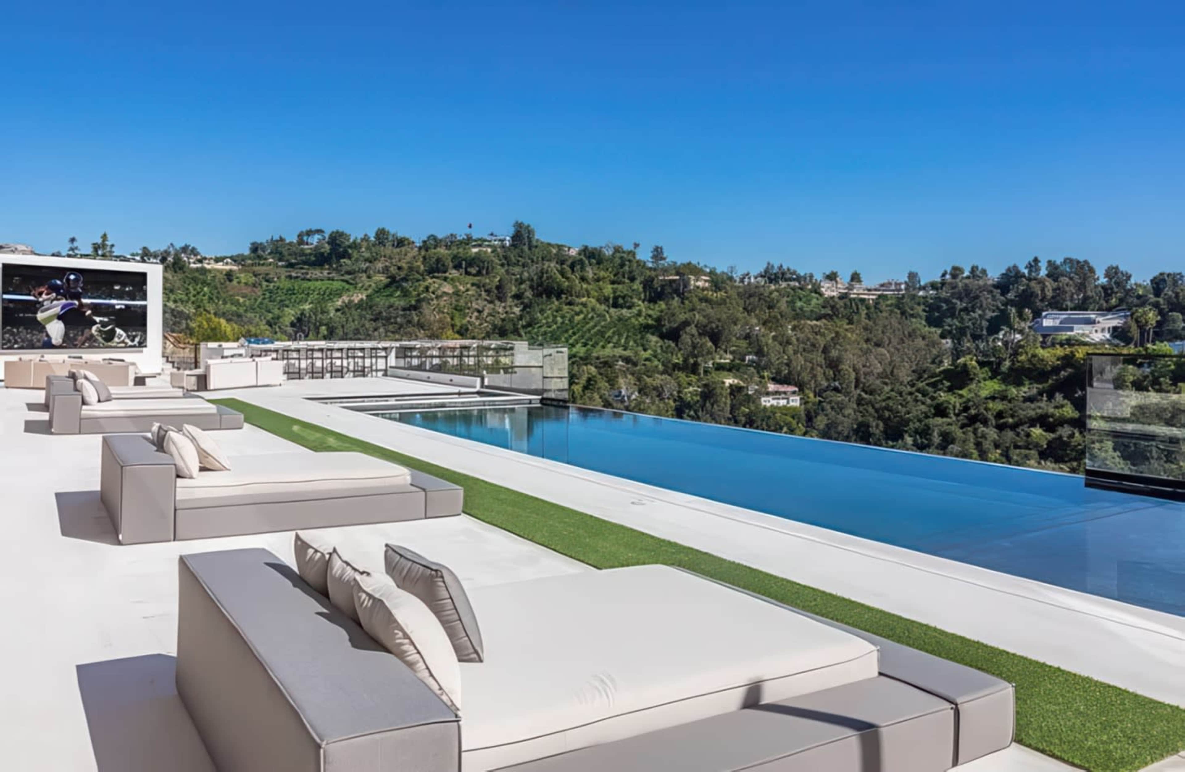 The image shows a luxurious outdoor terrace with modern lounge furniture, a swimming pool, and a scenic view of green hills under a clear blue sky.