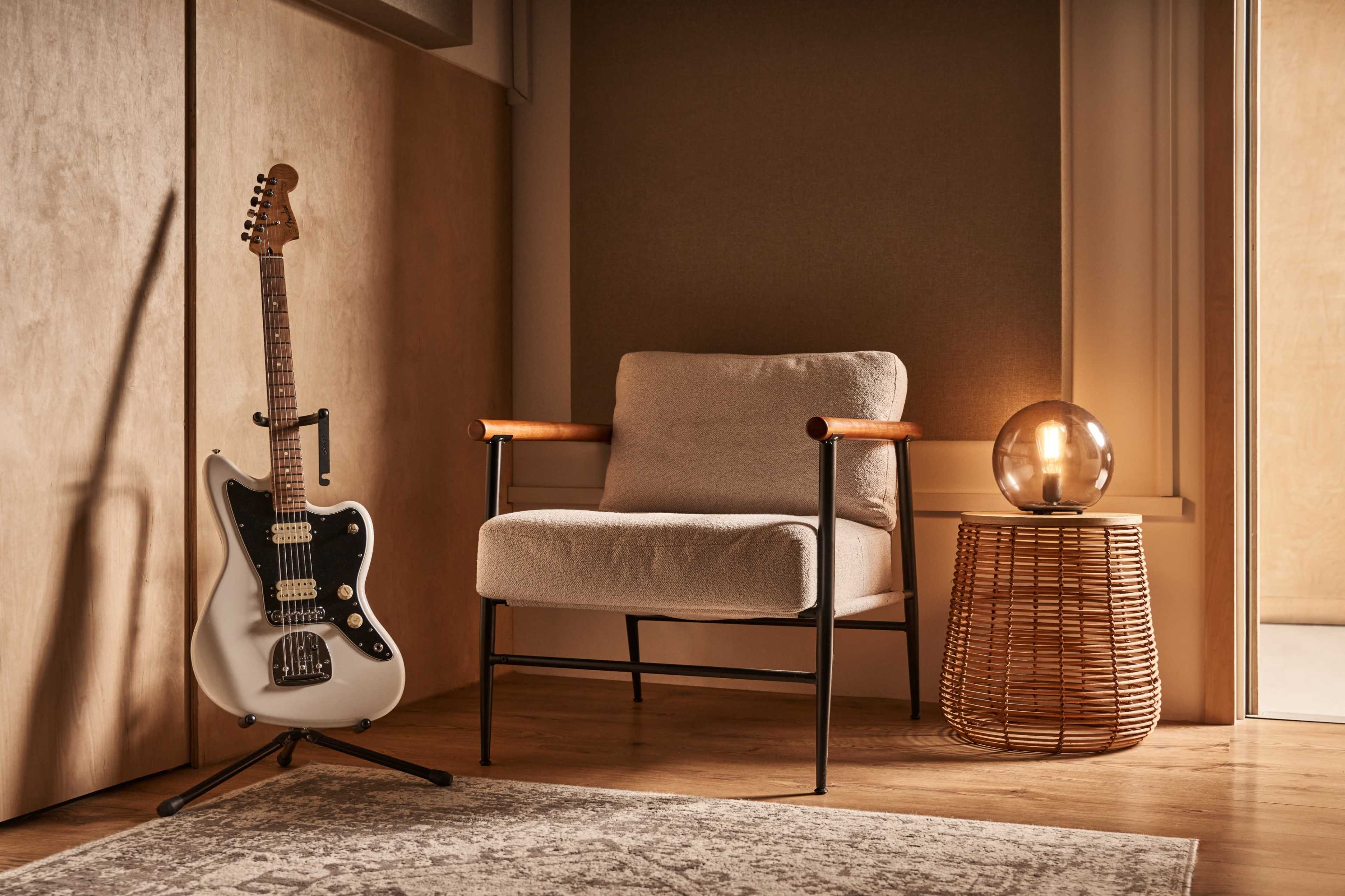 A white electric guitar stands in a corner next to a cozy armchair and a round woven lamp table.