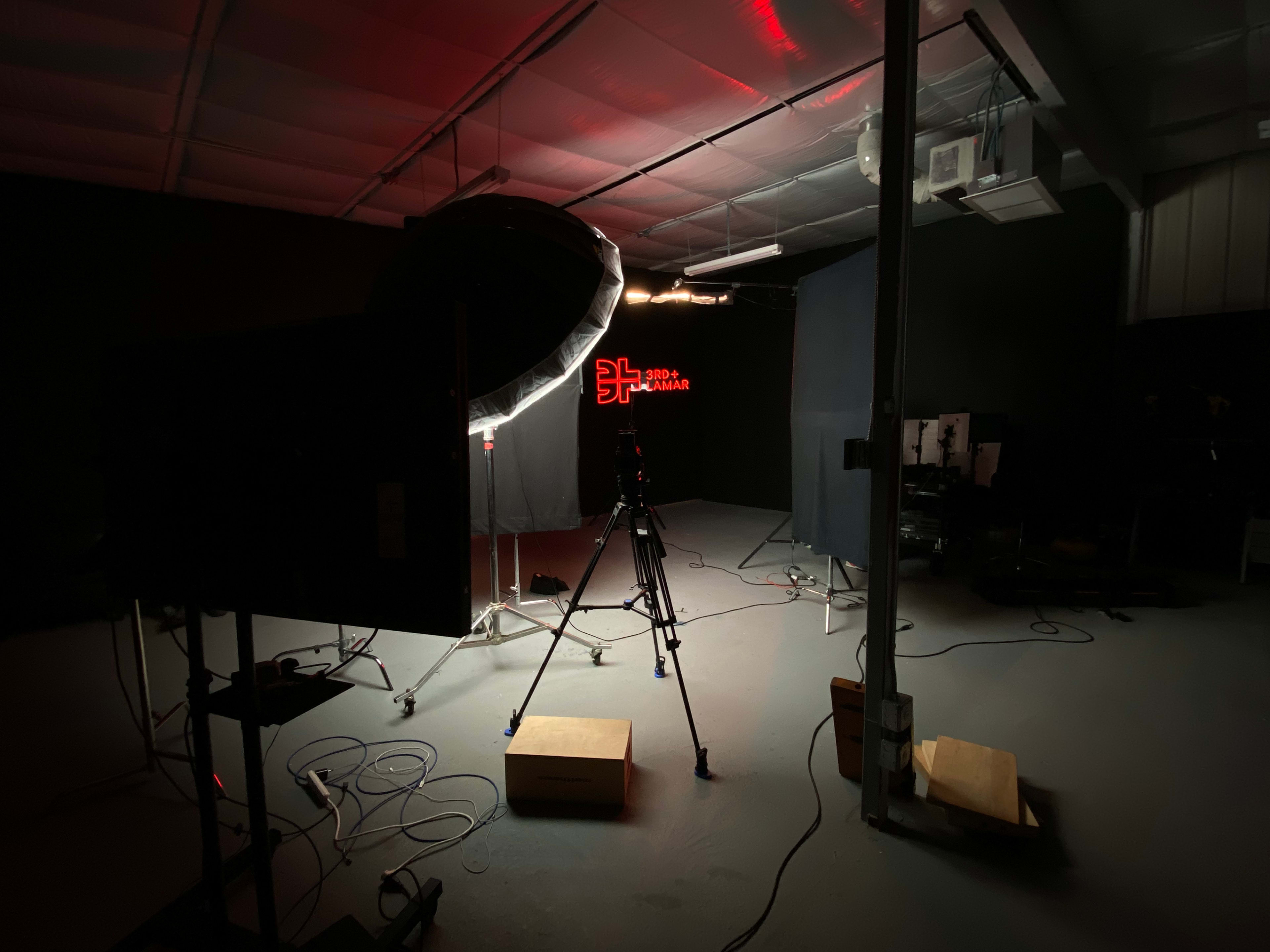 The image shows a dimly lit studio setup with a camera on a tripod, a large softbox light, and a background draped in black fabric.