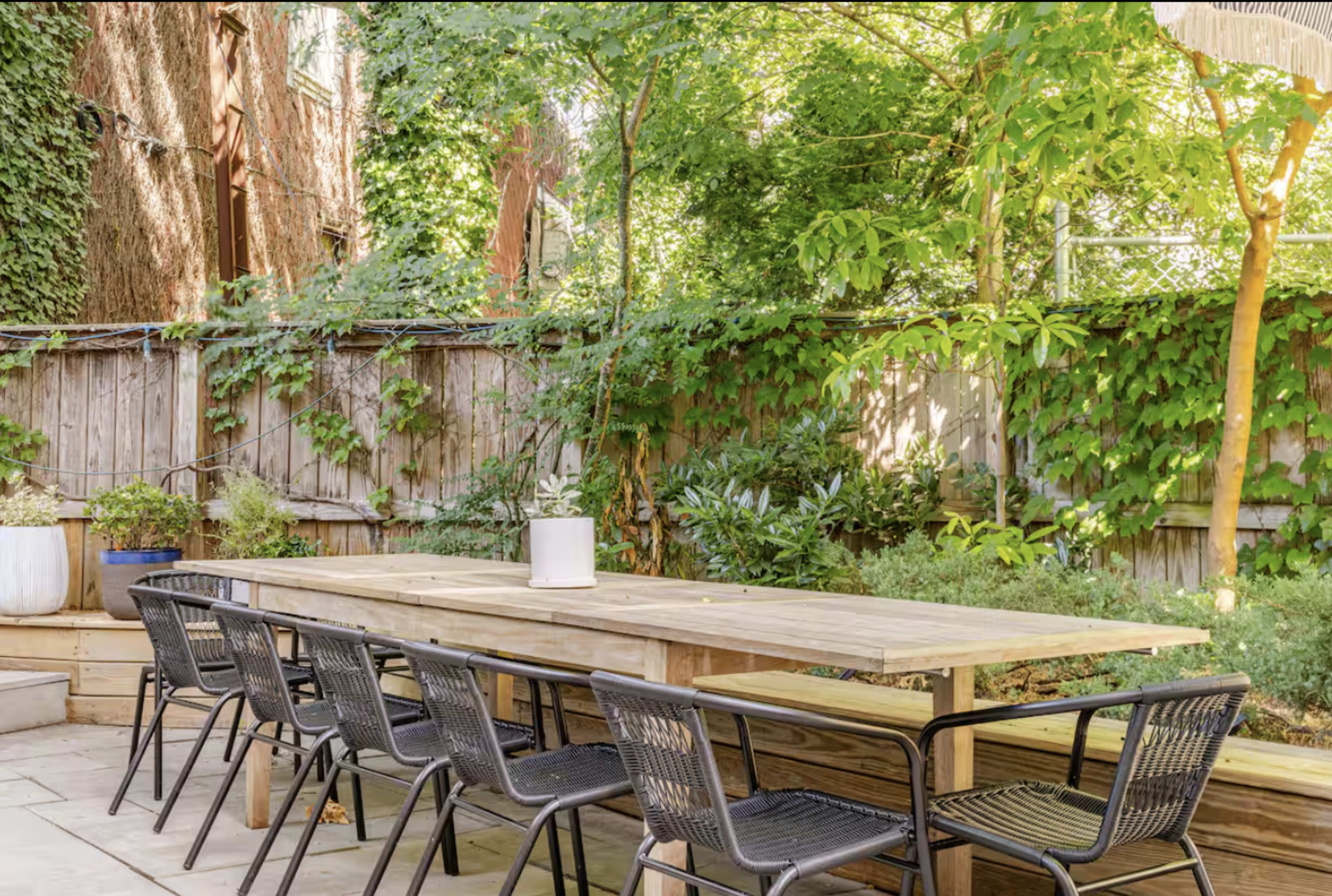 A long wooden dining table with black chairs is set in a green, leafy backyard surrounded by plants and a wooden fence.