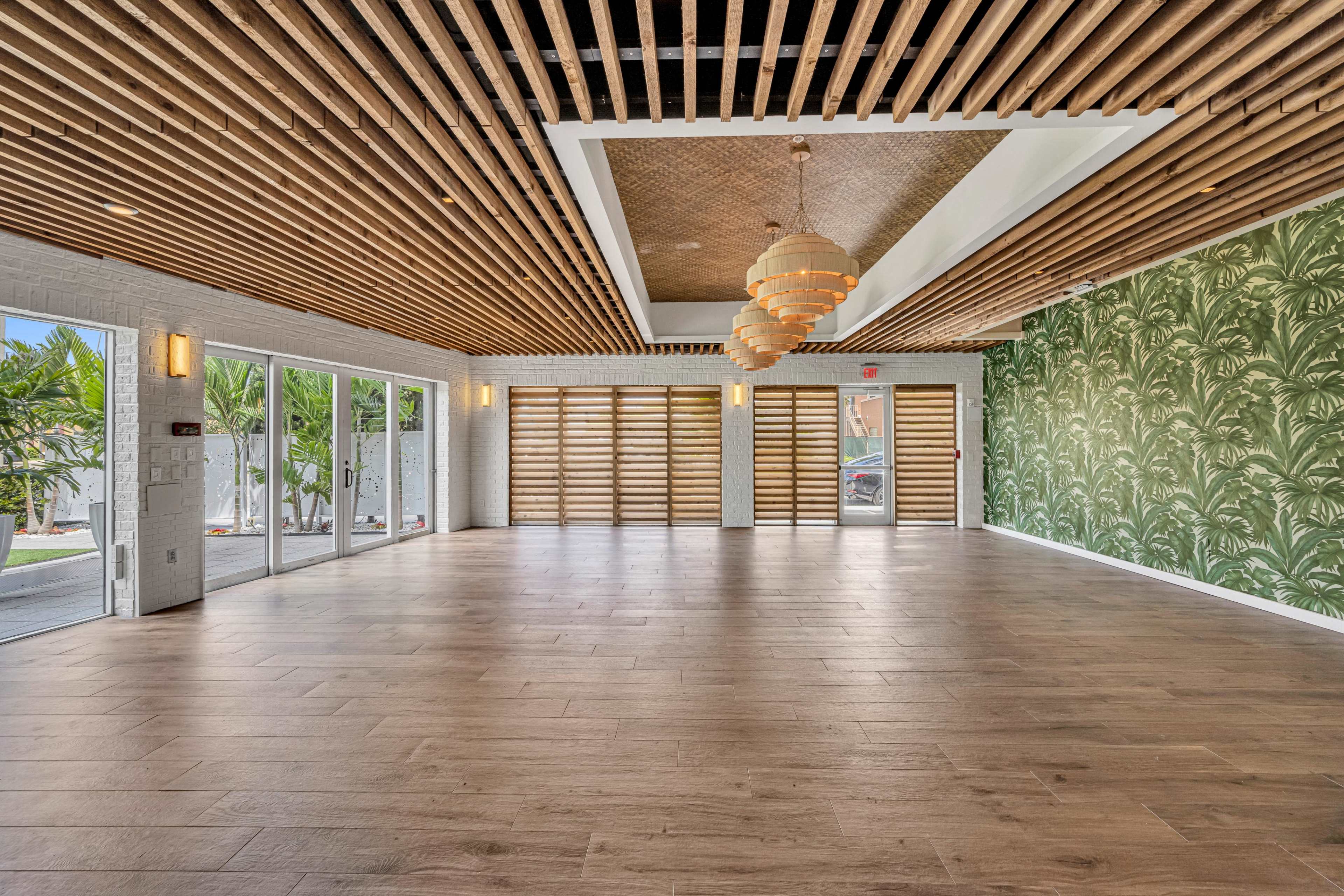 The image shows a spacious, modern interior room with wooden ceiling beams, a decorative light fixture, and one wall featuring a green palm leaf pattern.