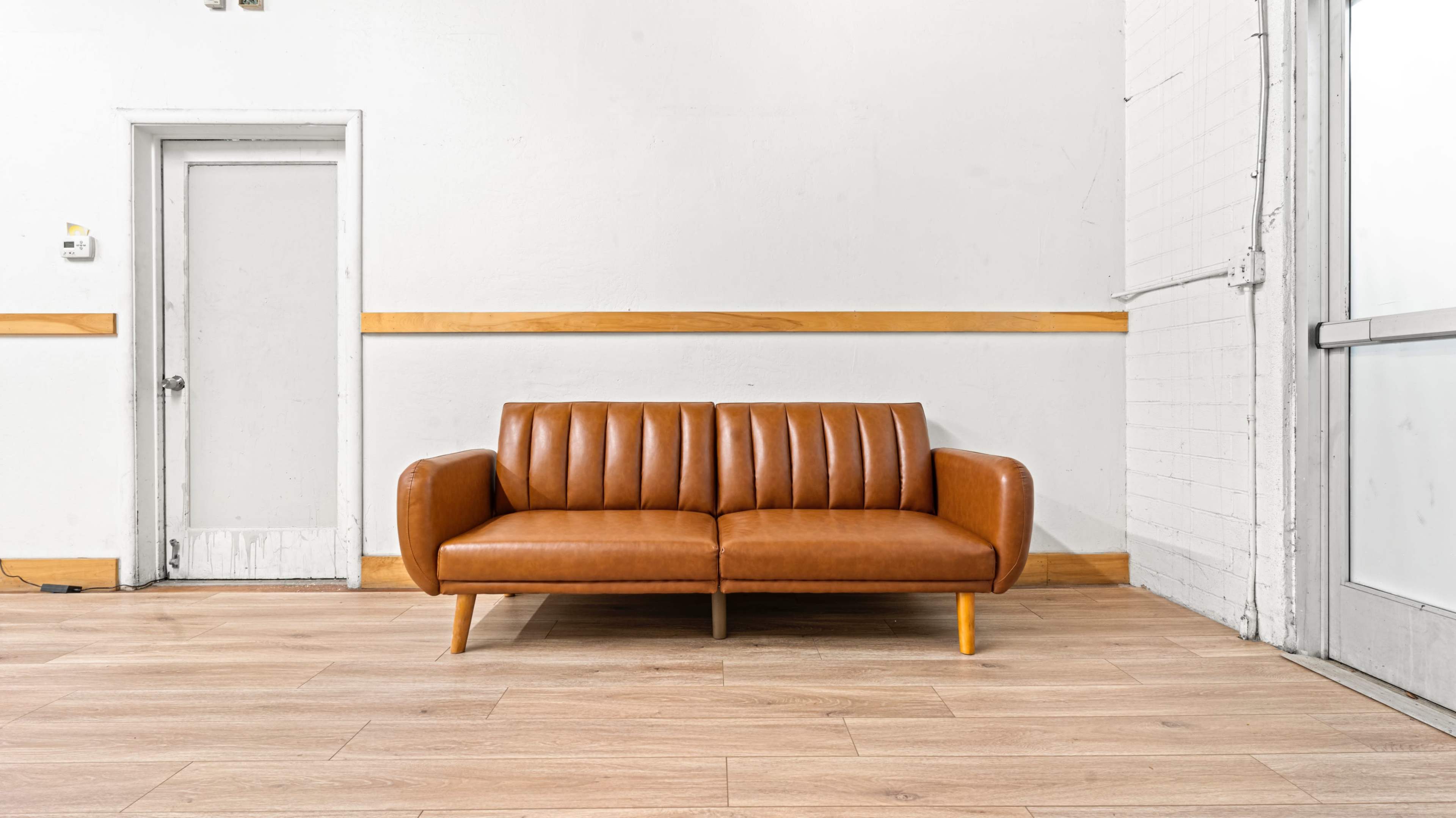A brown leather sofa with vertical stitching sits in a minimalist room with white walls and wooden flooring.