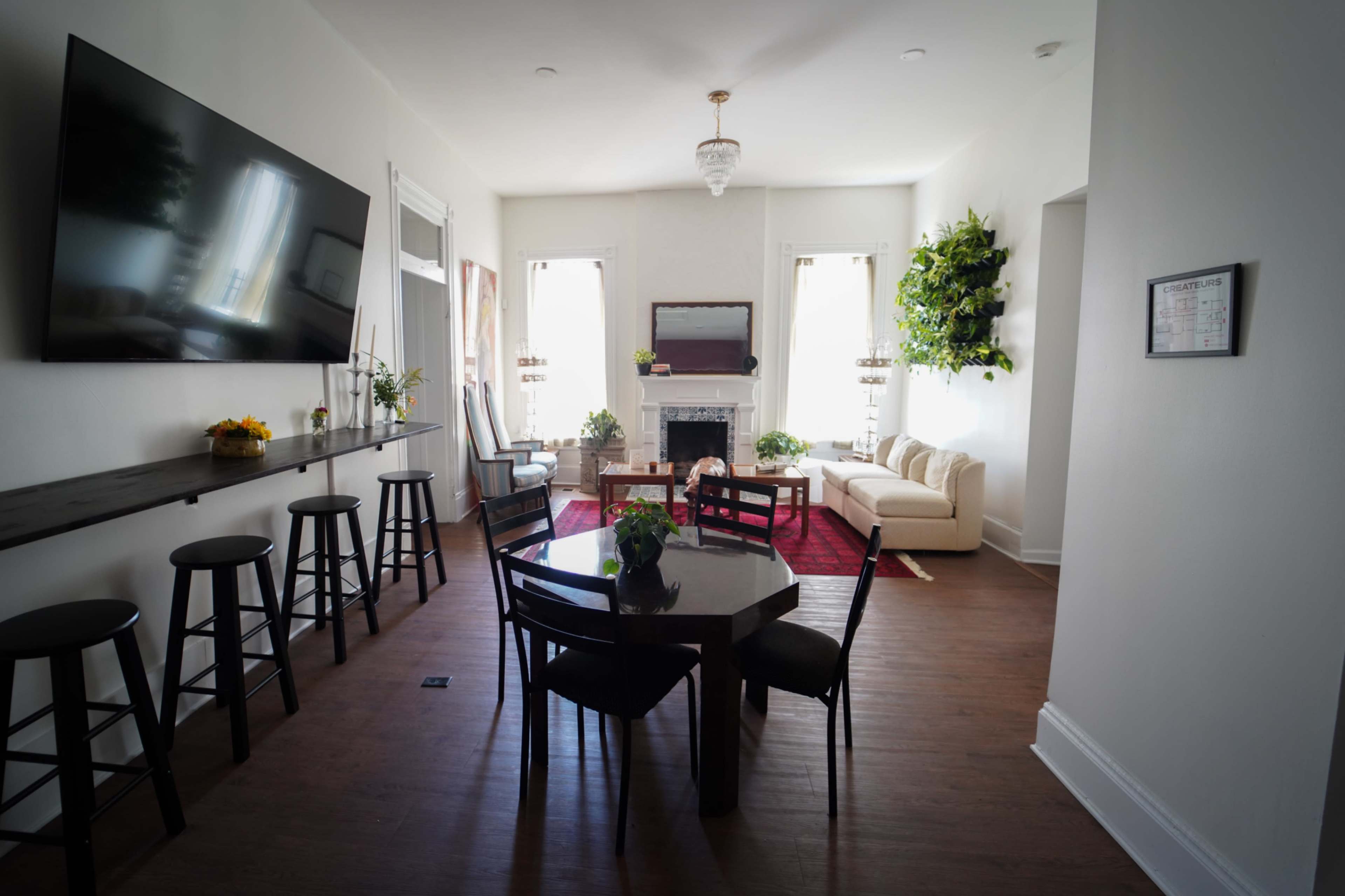 The image shows a bright living room with a dining area, featuring a round table and chairs, a sofa, and a large wall-mounted television.