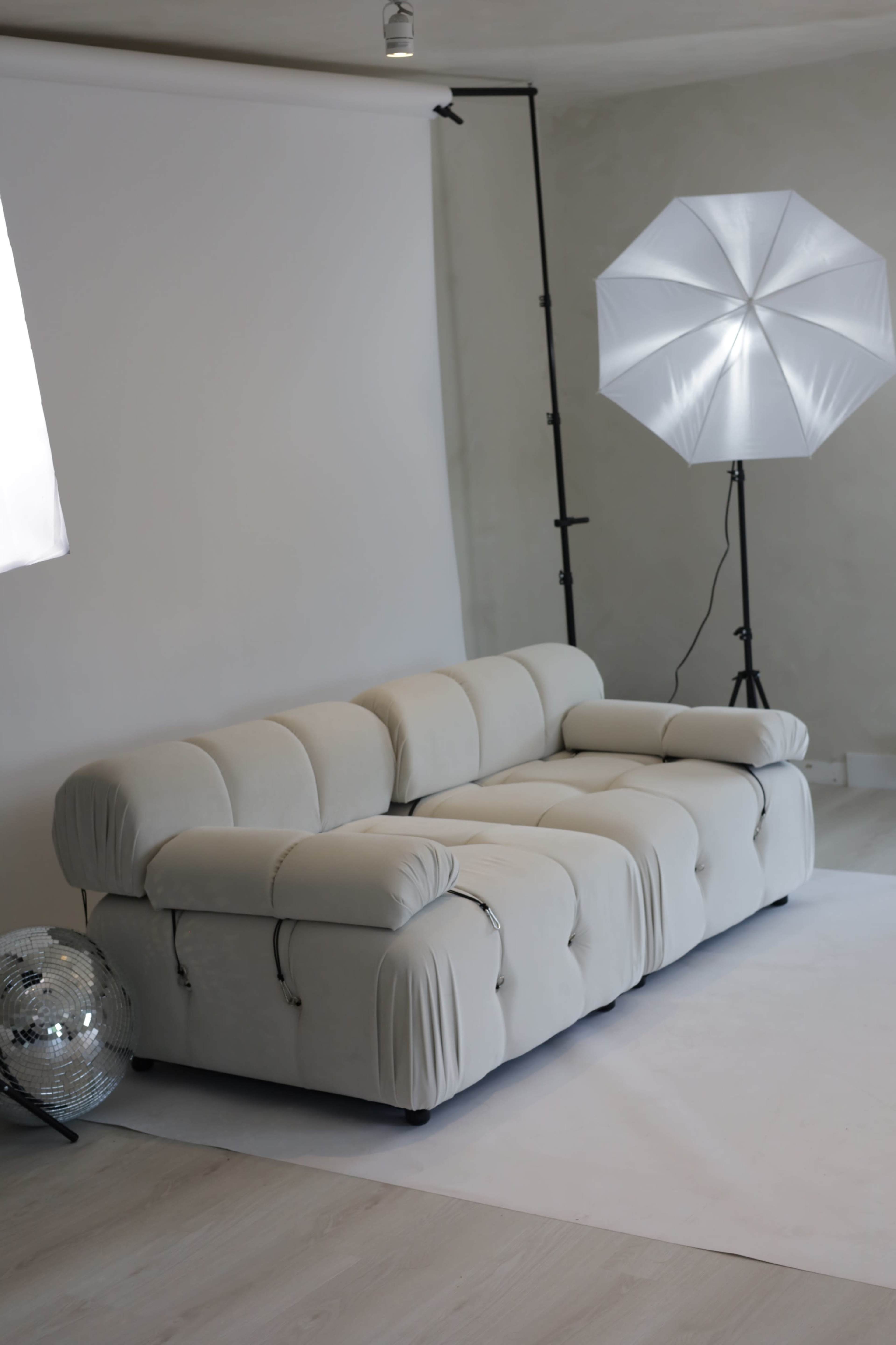 A beige sectional sofa is positioned in a minimalistic studio setting, accompanied by two softbox lights and a mirrored disco ball.