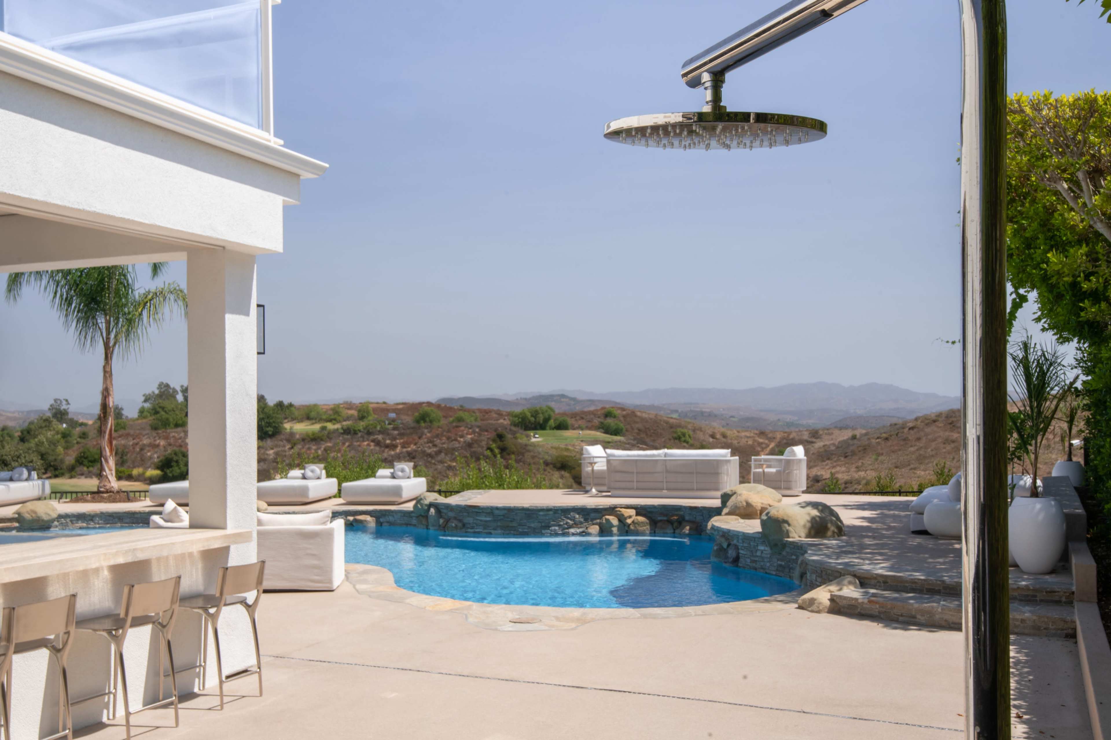 A modern outdoor space features a swimming pool surrounded by rock formations, lounge chairs, and a shower, with hills visible in the background.