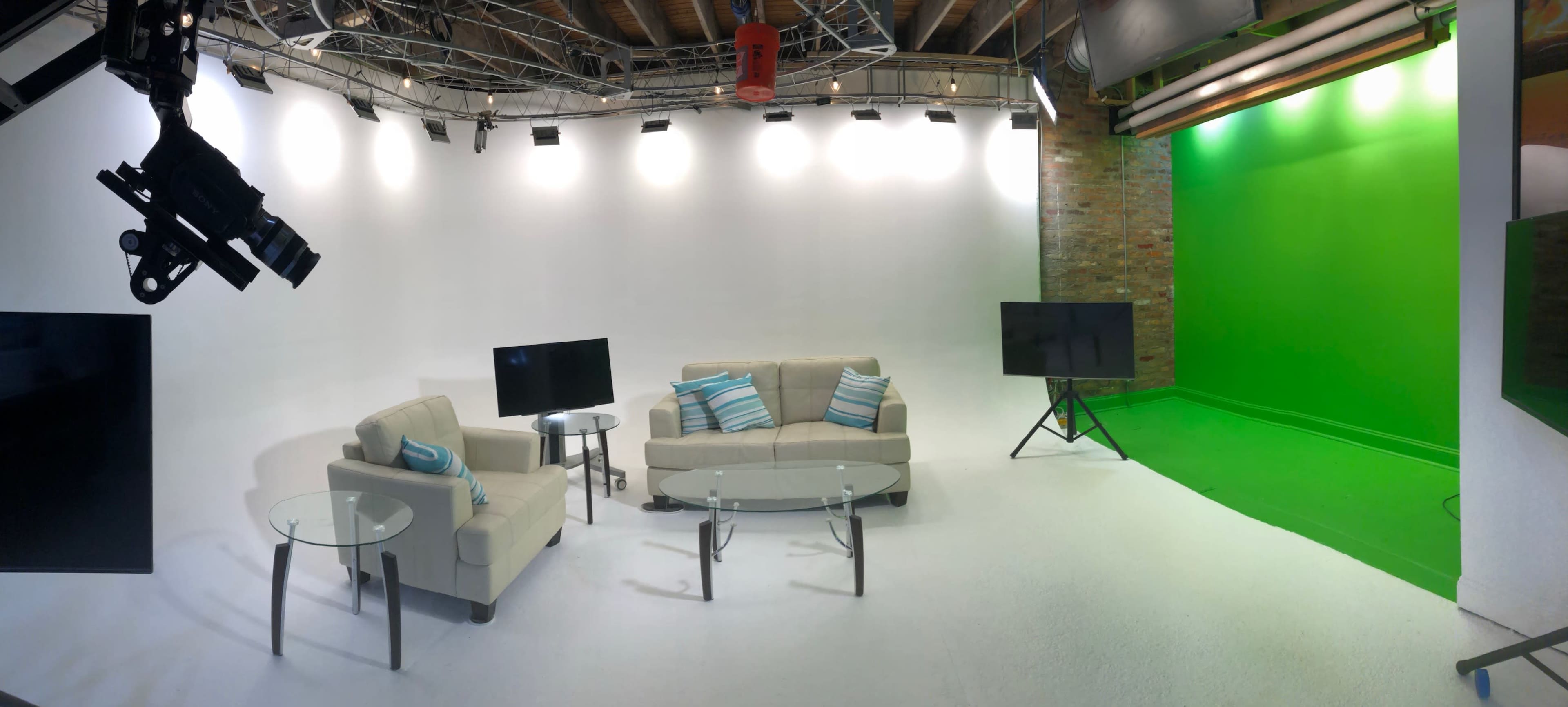A studio setup with two light-colored sofas, glass coffee tables, and two television screens in front of a green screen backdrop.