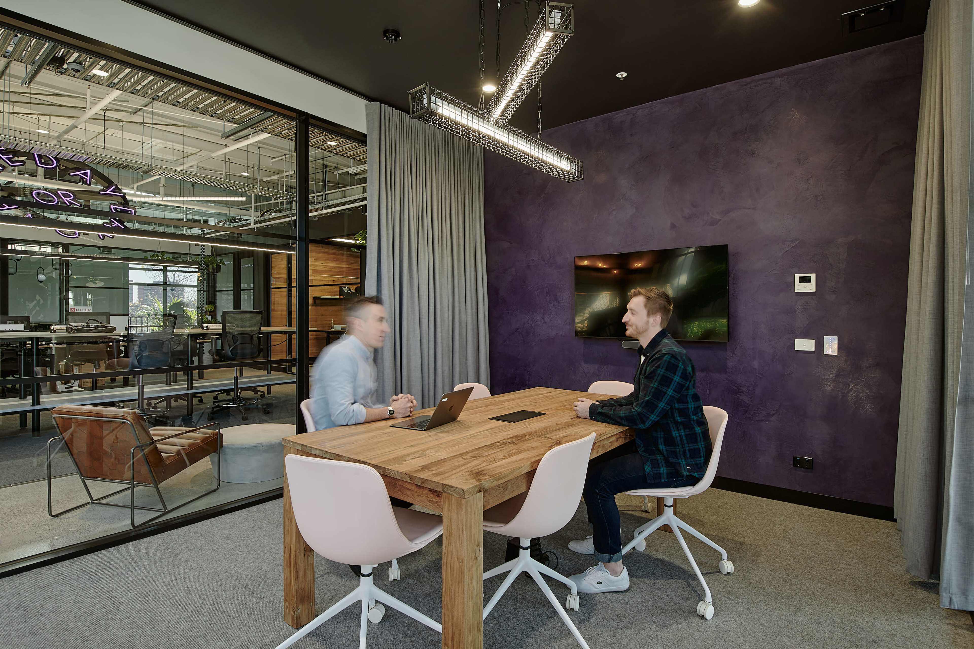 Two people are seated at a wooden conference table in a modern office space with a dark wall and a television display.