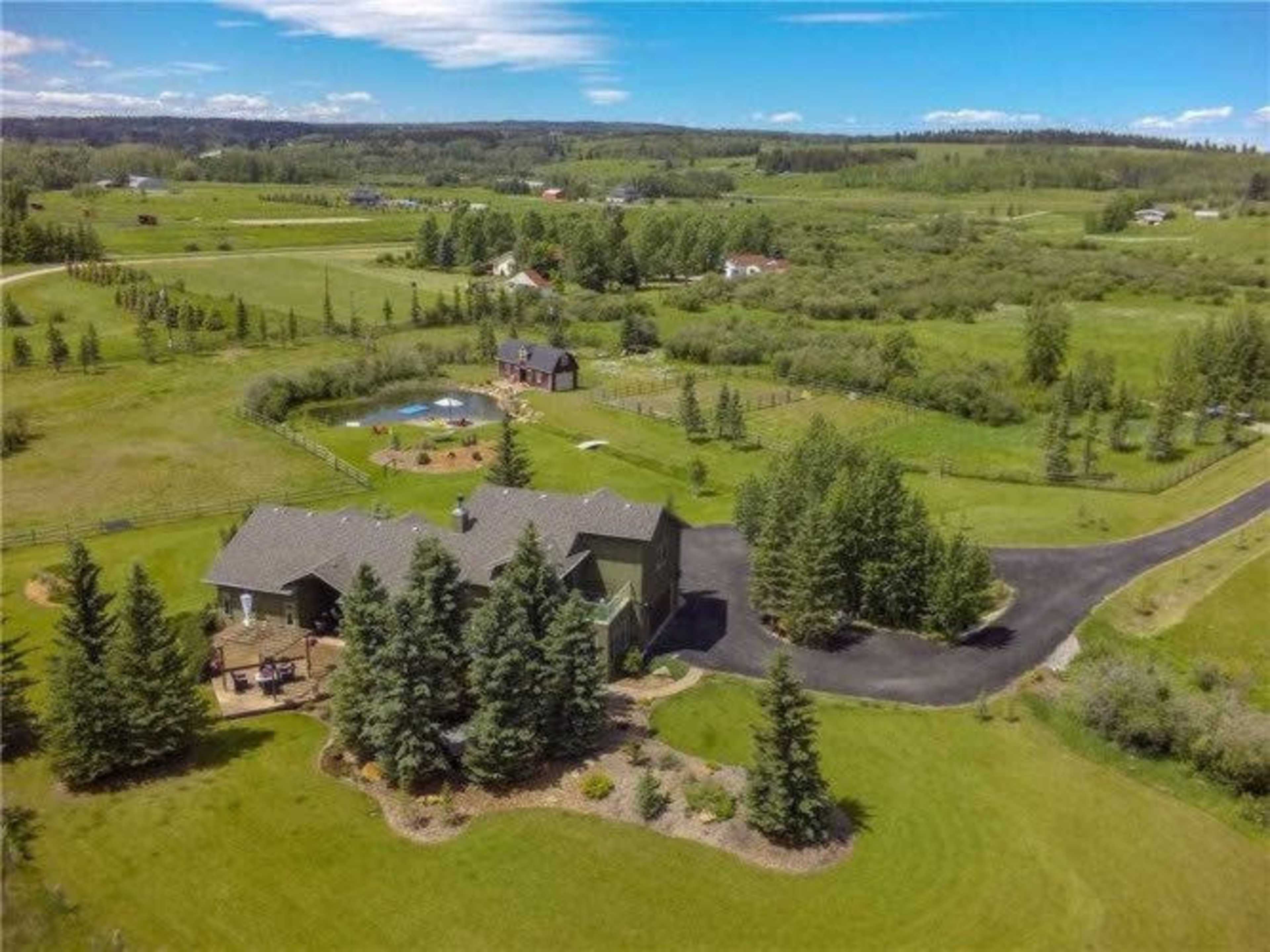 The image shows a sprawling green landscape with a house in the foreground surrounded by trees and a winding driveway, while a smaller building and a pool are visible in the background.