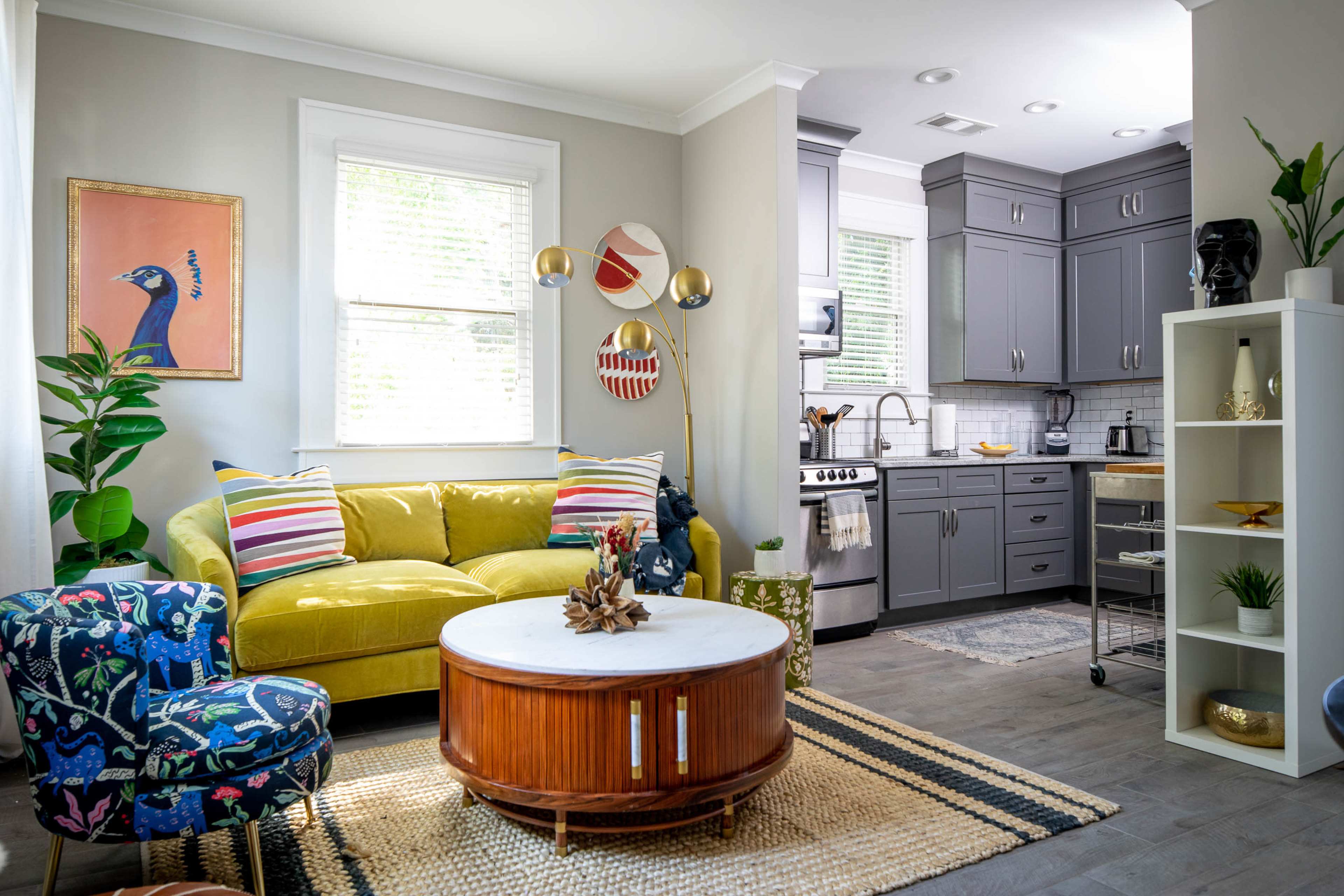 The image shows a stylish living room with a green couch, colorful chairs, and a round coffee table, adjacent to a modern kitchen with grey cabinets and stainless steel appliances.