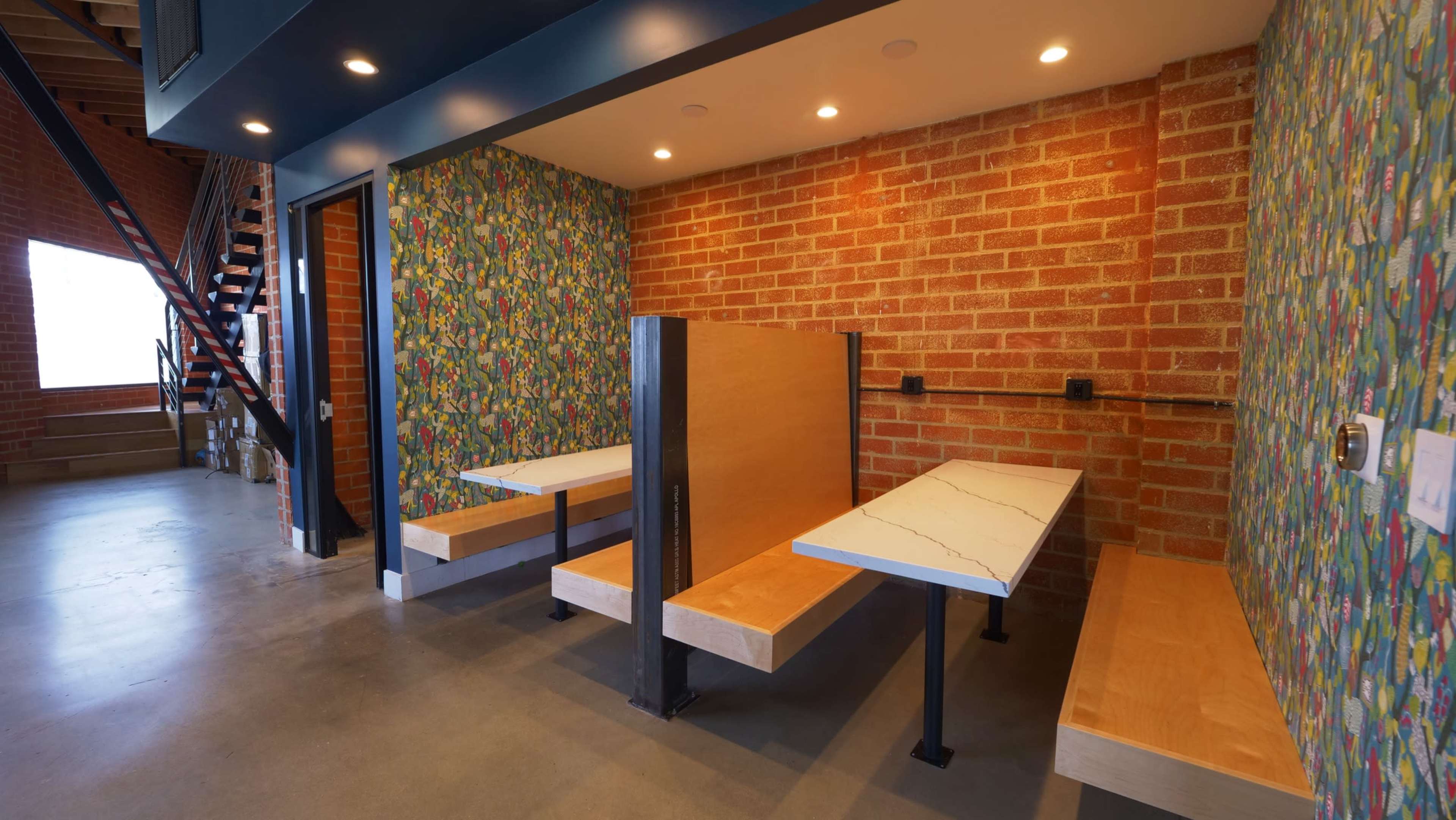 The image shows a modern interior with wooden benches and tables along a colorful, patterned wall in a space with exposed brick.