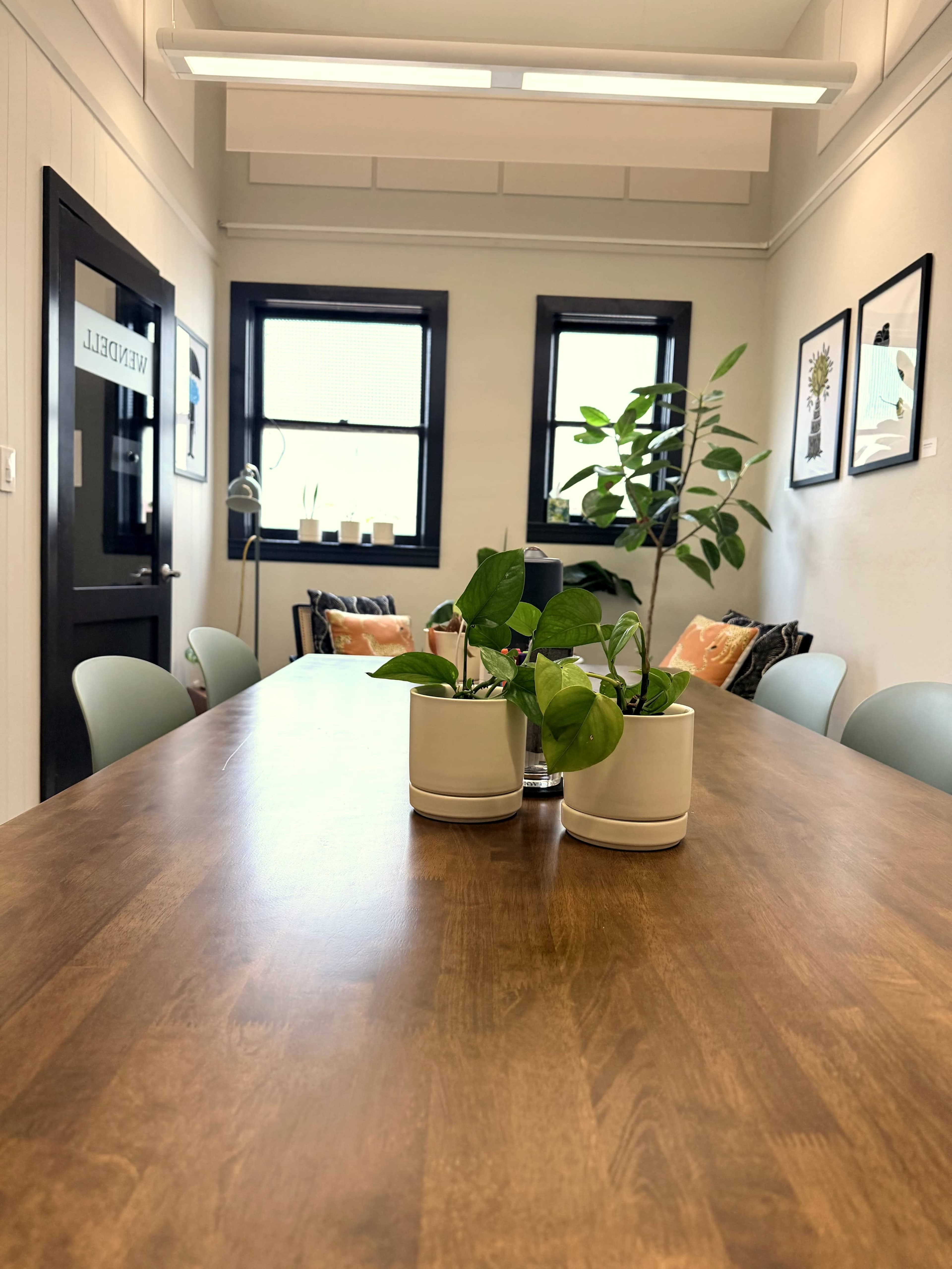 A wooden conference table with potted plants is centered in a brightly lit room decorated with artwork on the walls and chairs positioned around the table.