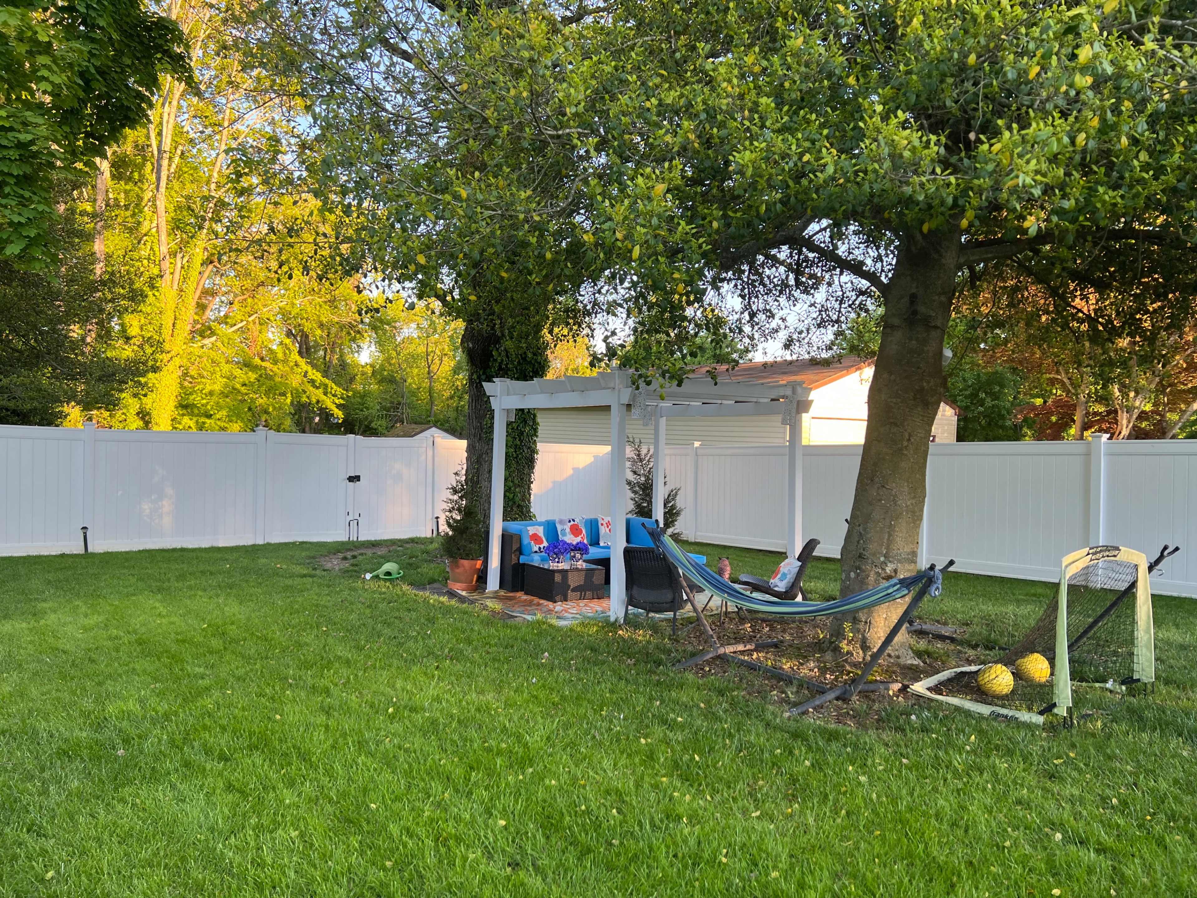 A grassy backyard features a hammock suspended from a tree, a small fire pit area, and a white fence surrounding the space.