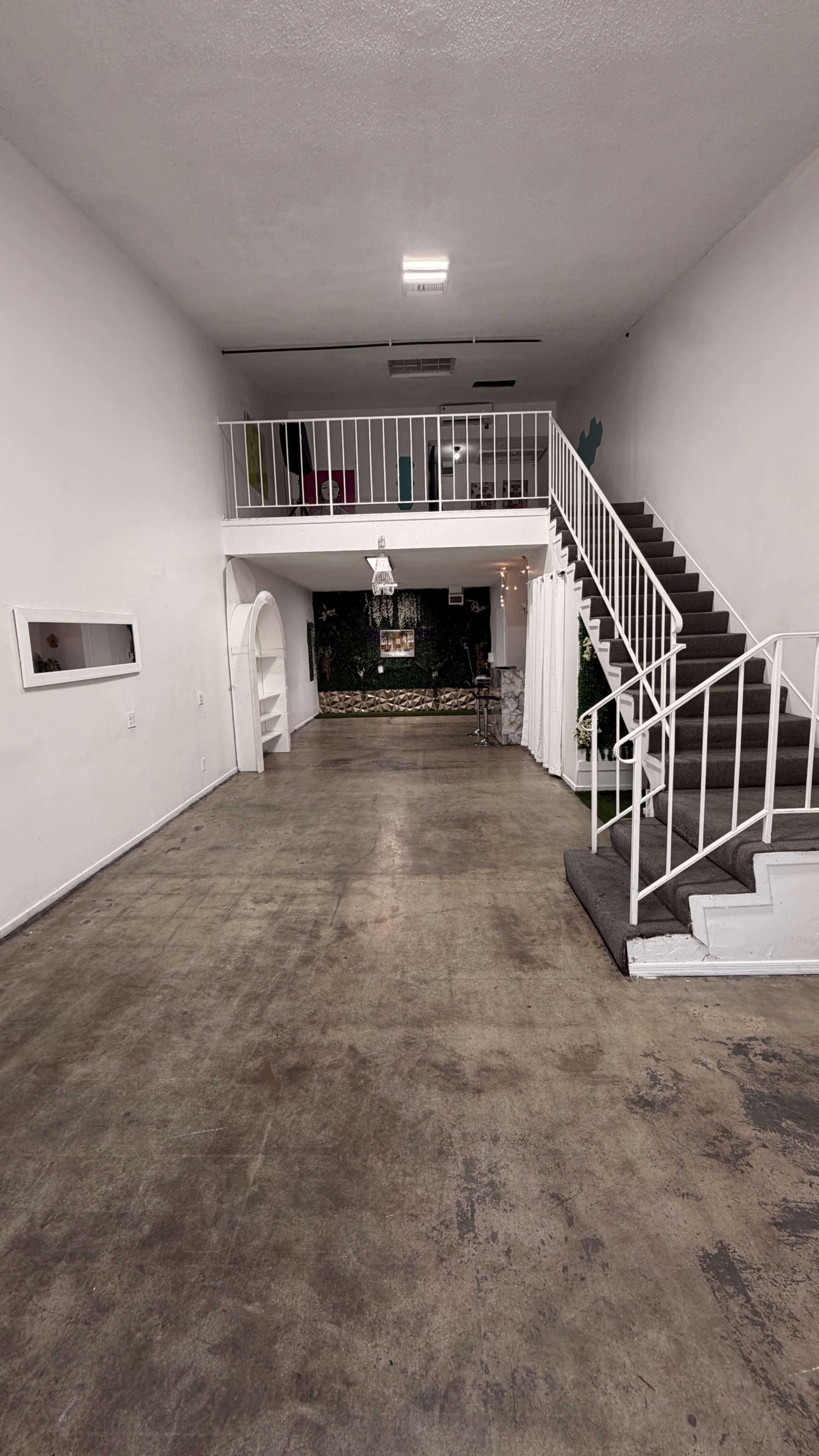 The image shows a spacious, empty room with a staircase on the right leading to an upper level, and a small area with decorative plants in the background.