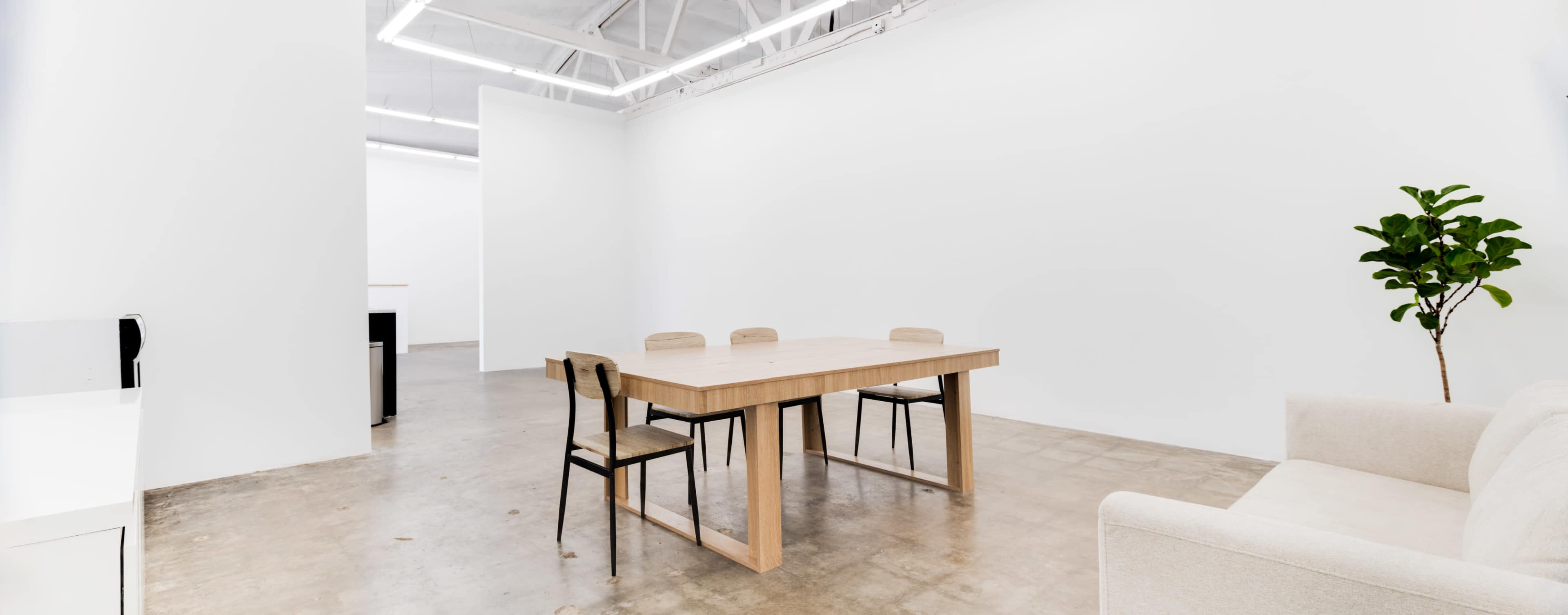A minimalistic office space features a wooden table surrounded by chairs, a couch, and a potted plant against white walls.