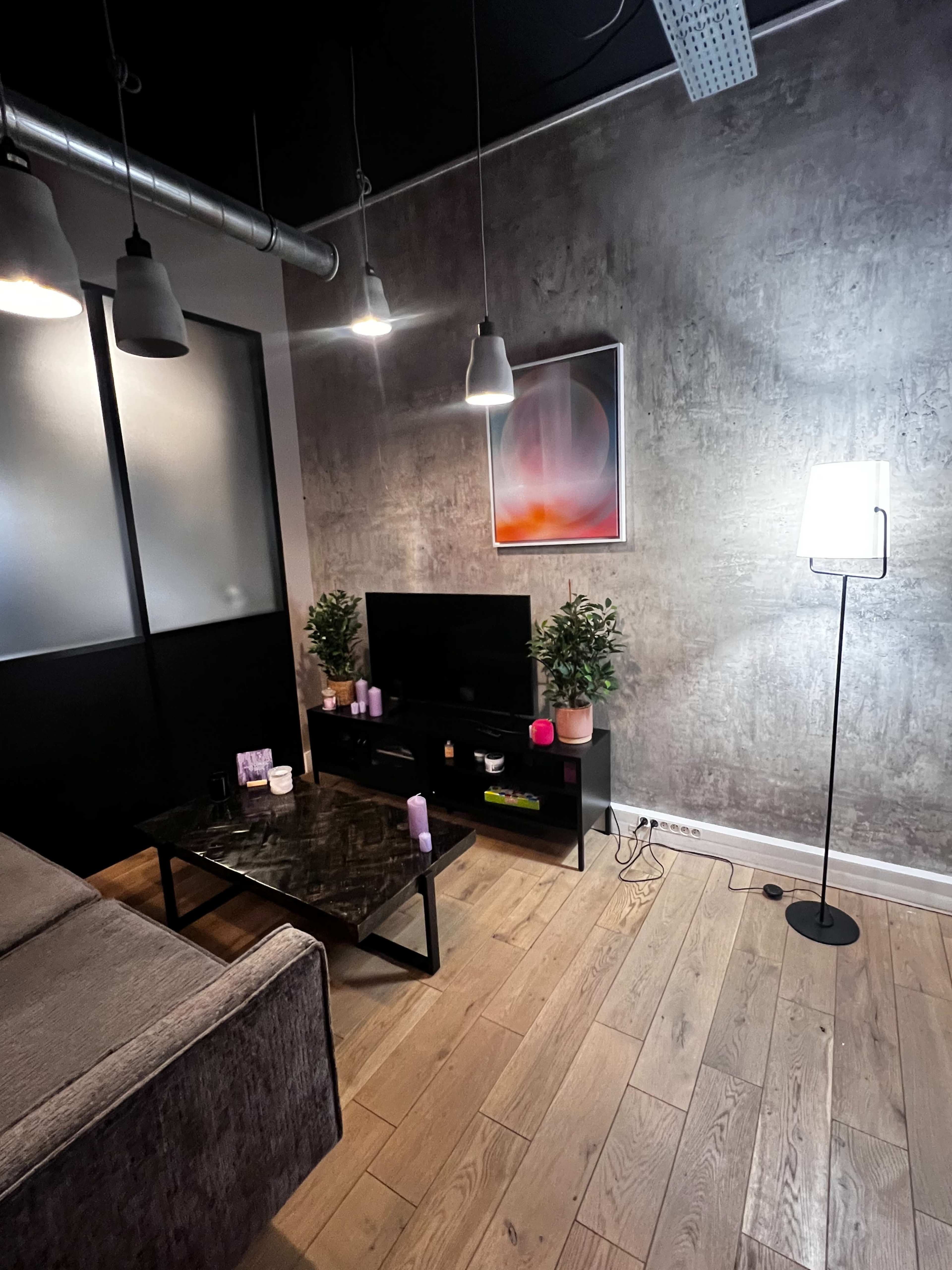 The image shows a modern living room with a gray wall, a television with decorative items on a black media unit, a gray sofa, and ambient lighting from pendant lamps and a floor lamp.