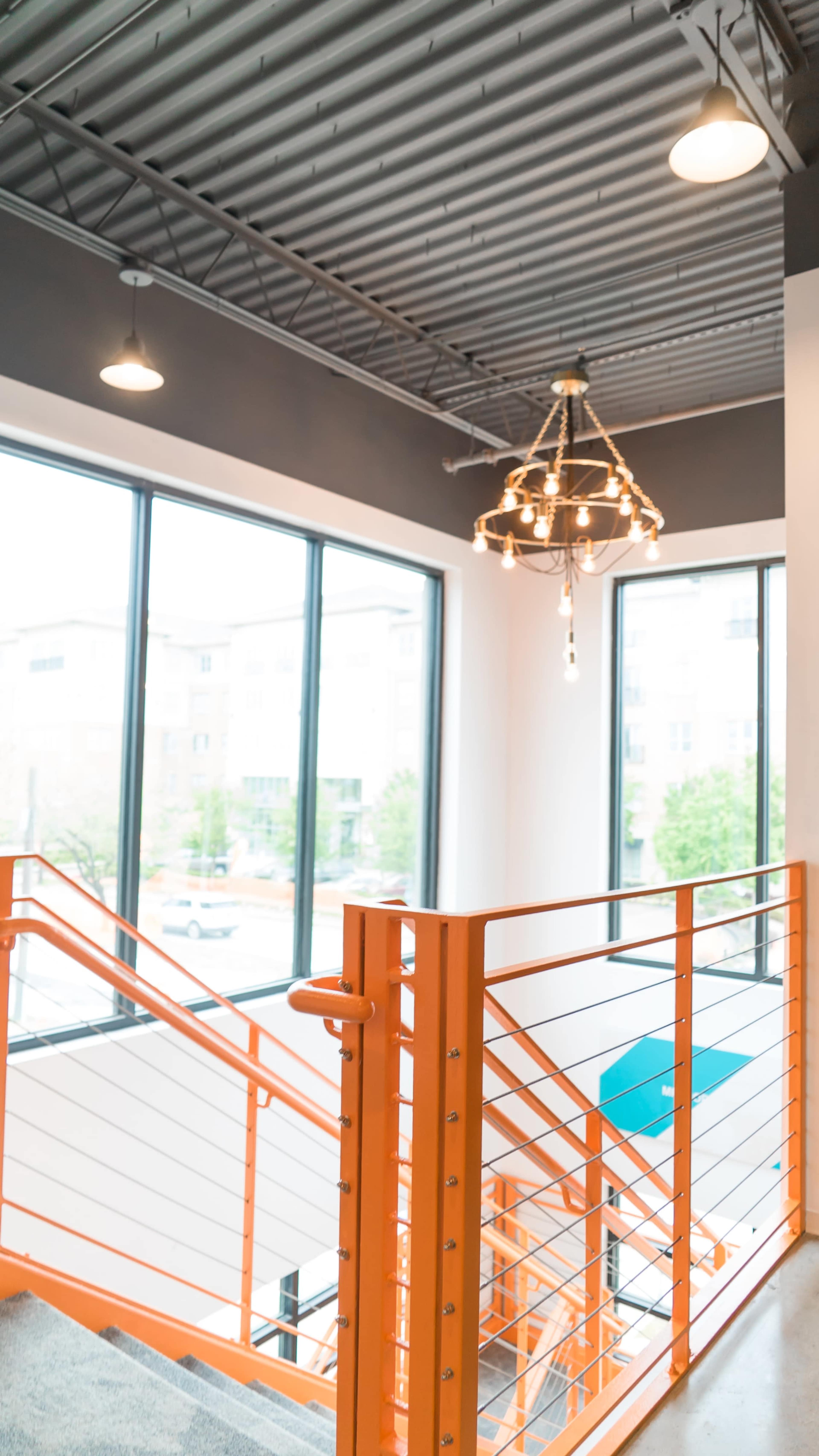 The image shows a brightly lit staircase with an orange railing, large windows, and an industrial-style light fixture hanging from the ceiling.