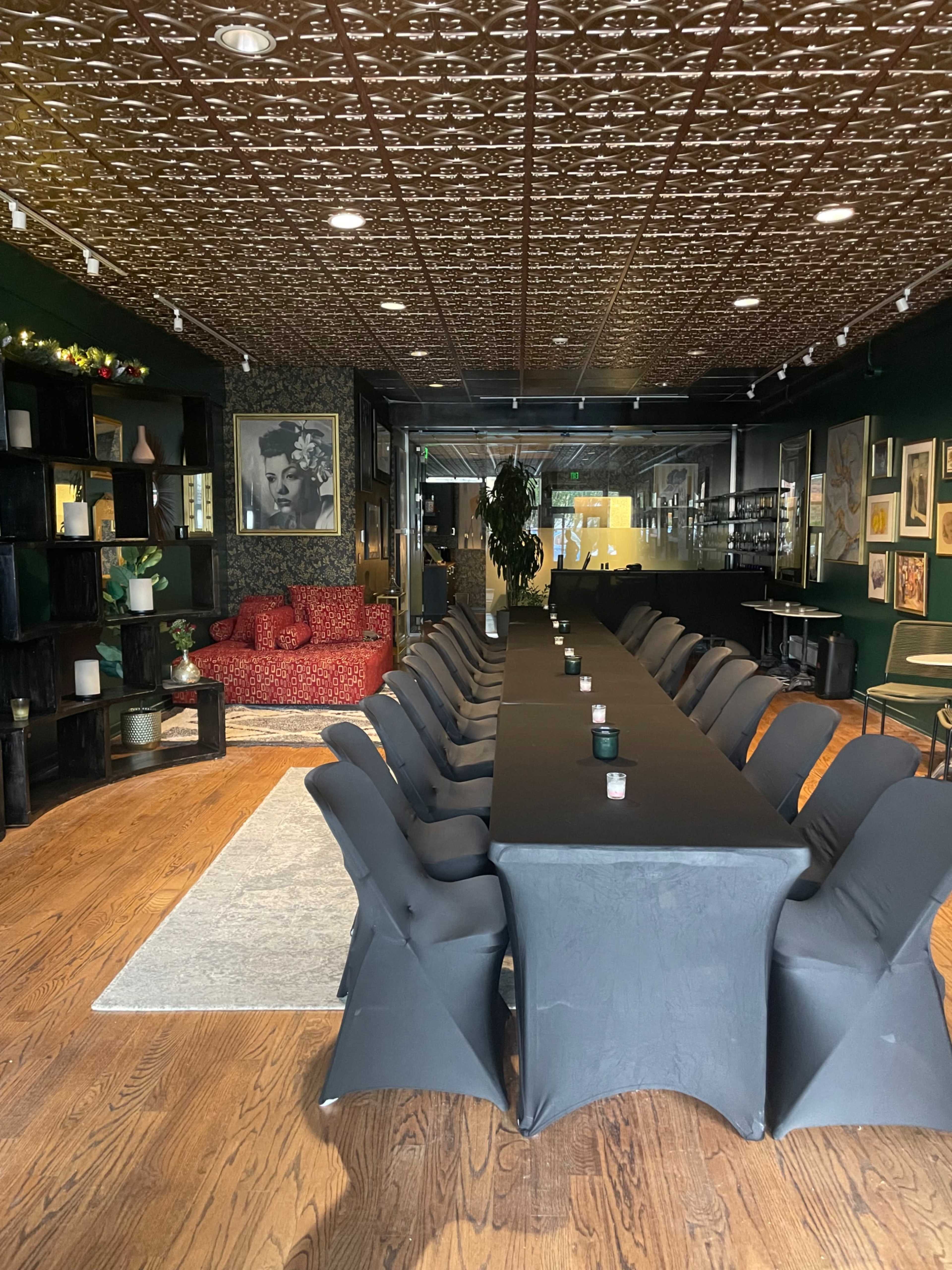 The image shows a long table set for a gathering in a dimly lit room with dark green walls, a decorative ceiling, and eclectic artwork on the walls.