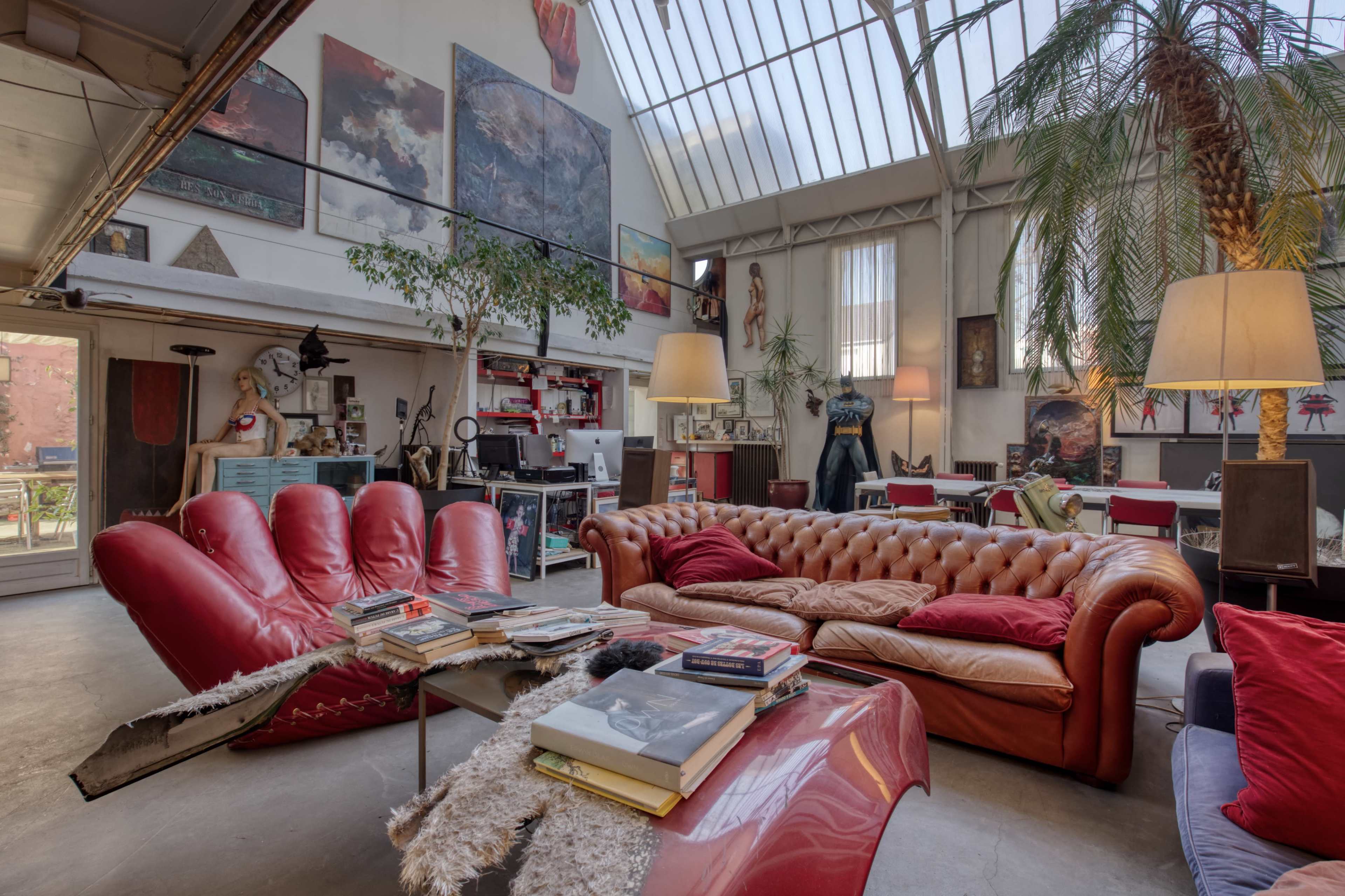 A spacious, eclectic living room featuring a red chesterfield sofa, a glass coffee table piled with books, and various artwork on the walls.