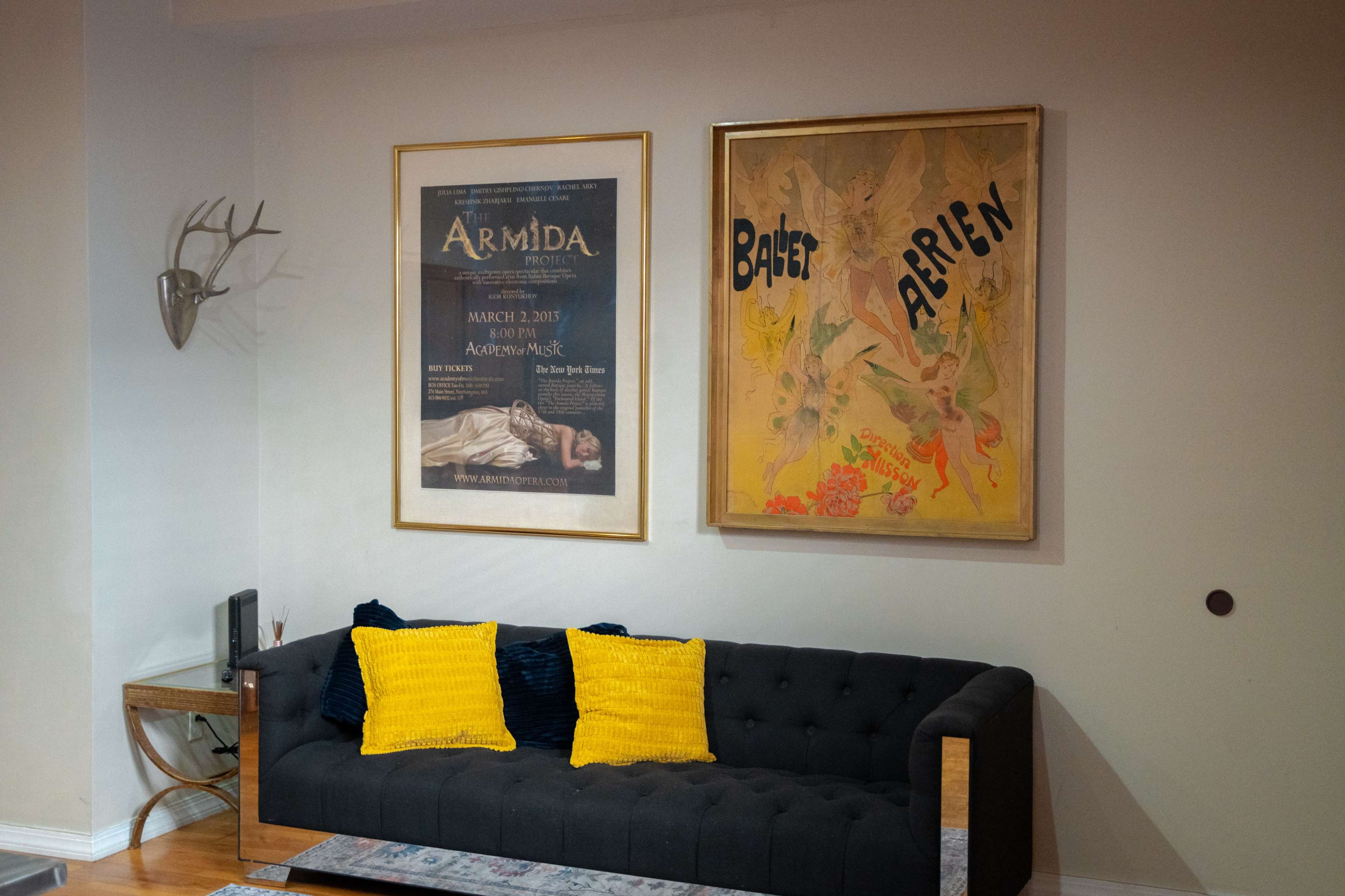The image shows a living room with a dark sofa adorned with yellow throw pillows, flanked by two framed posters on the walls.
