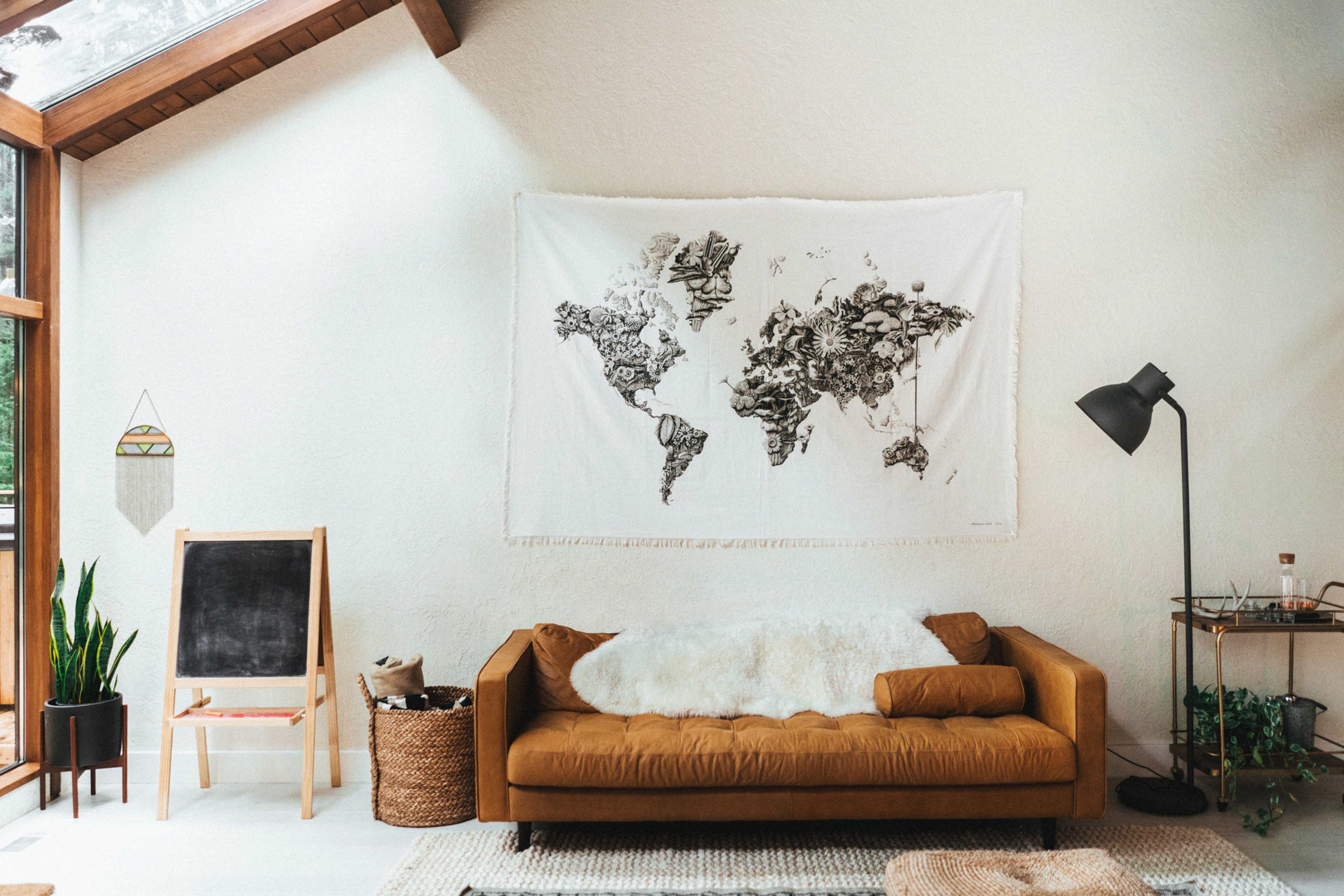 A world map tapestry hangs on a white wall above a brown couch in a room with large windows, a chalkboard, a plant, and a floor lamp.