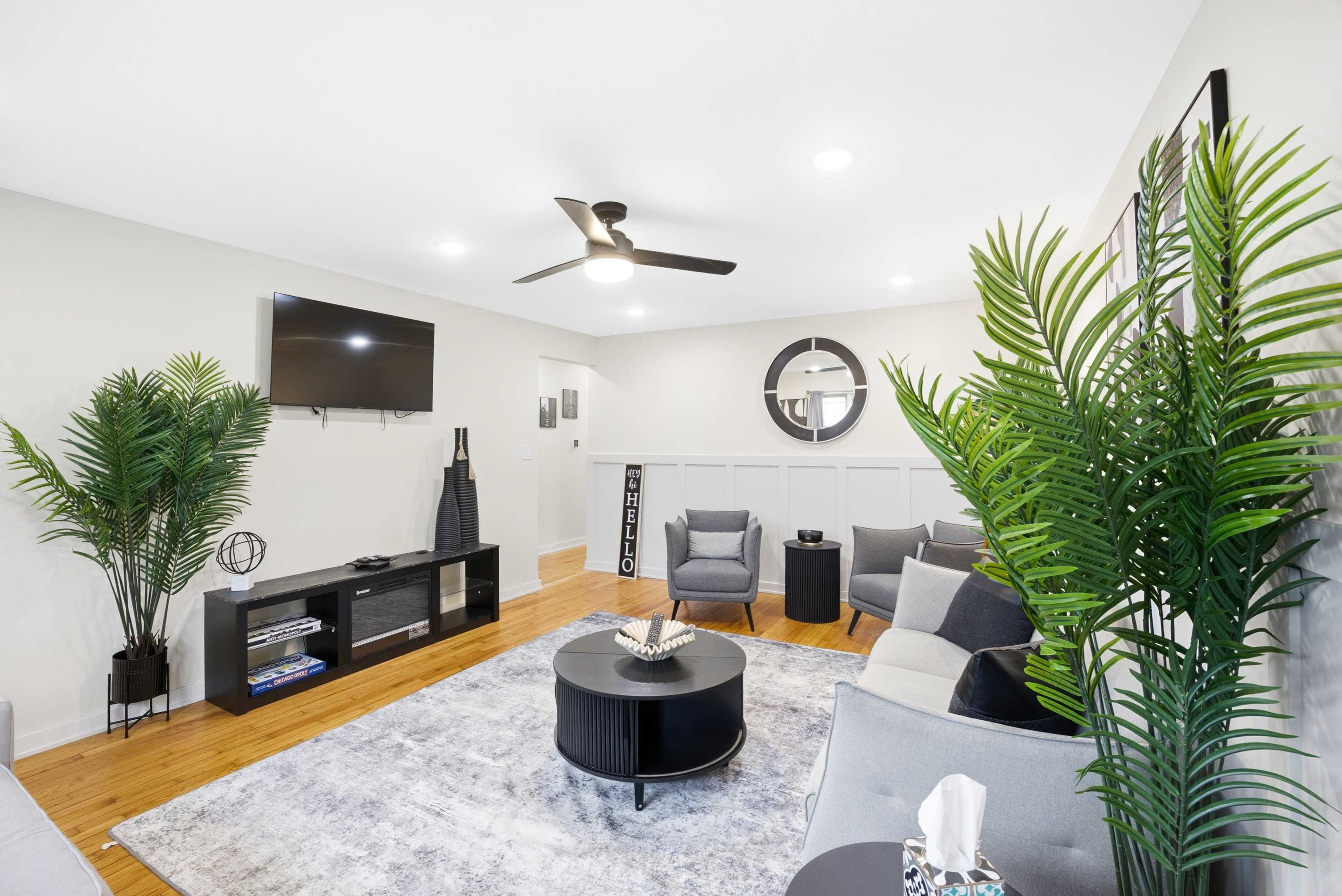 A modern living room features two gray armchairs, a black coffee table, and a television mounted on the wall, surrounded by decorative plants and a light-colored area rug.