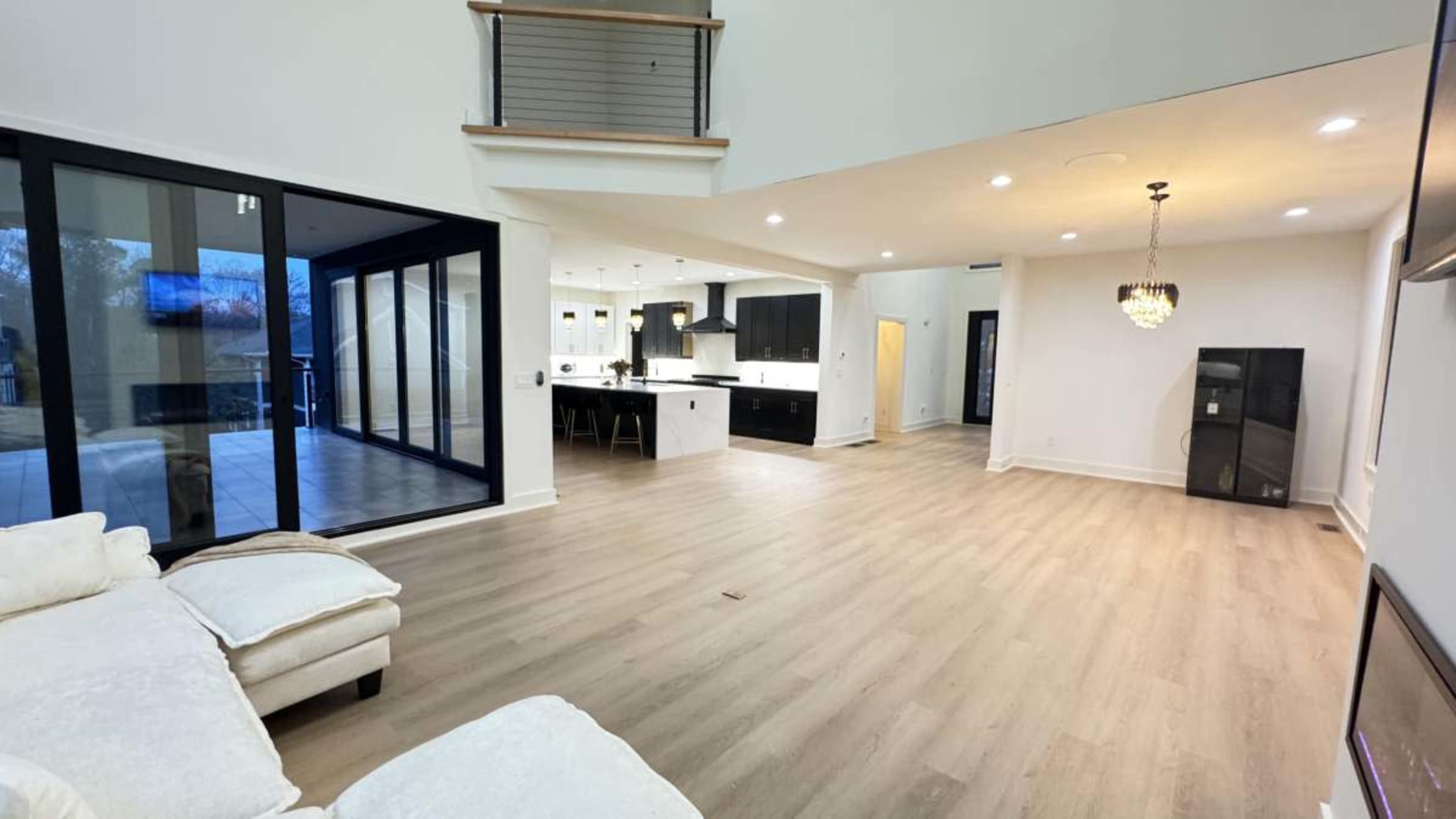 The image shows a spacious, modern living area with a light wood floor, a sectional sofa, and large windows leading to an outdoor space.