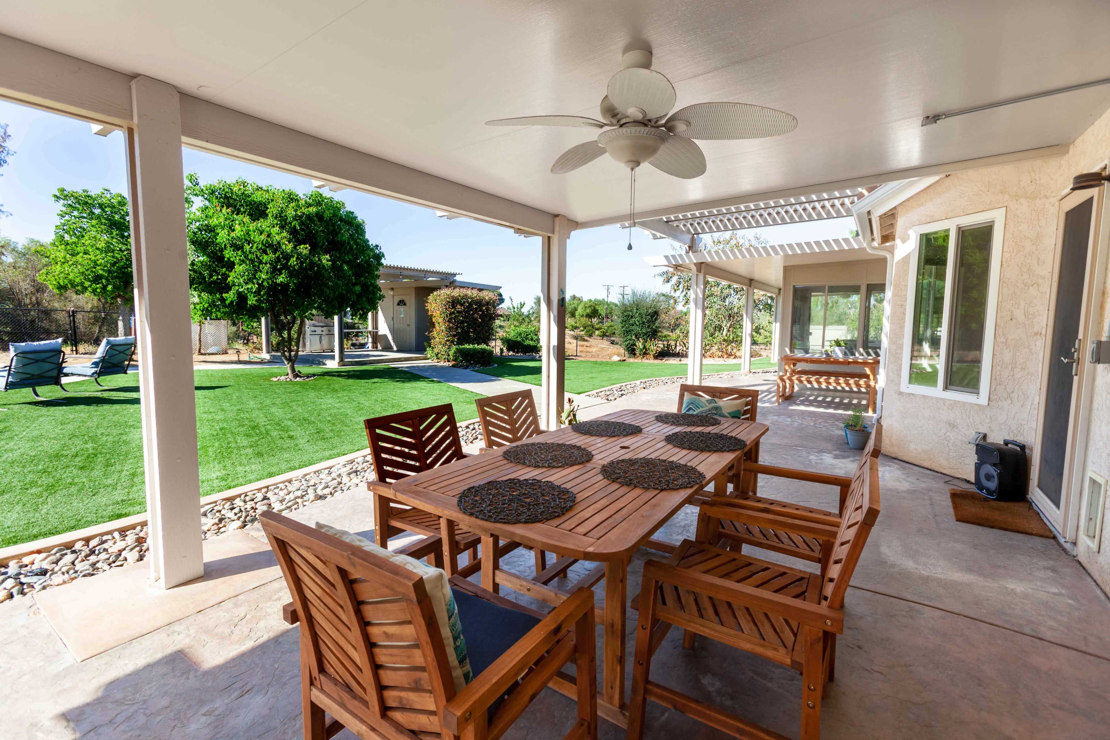 A wooden dining set is arranged under a covered patio, overlooking a lawn and landscaped yard.