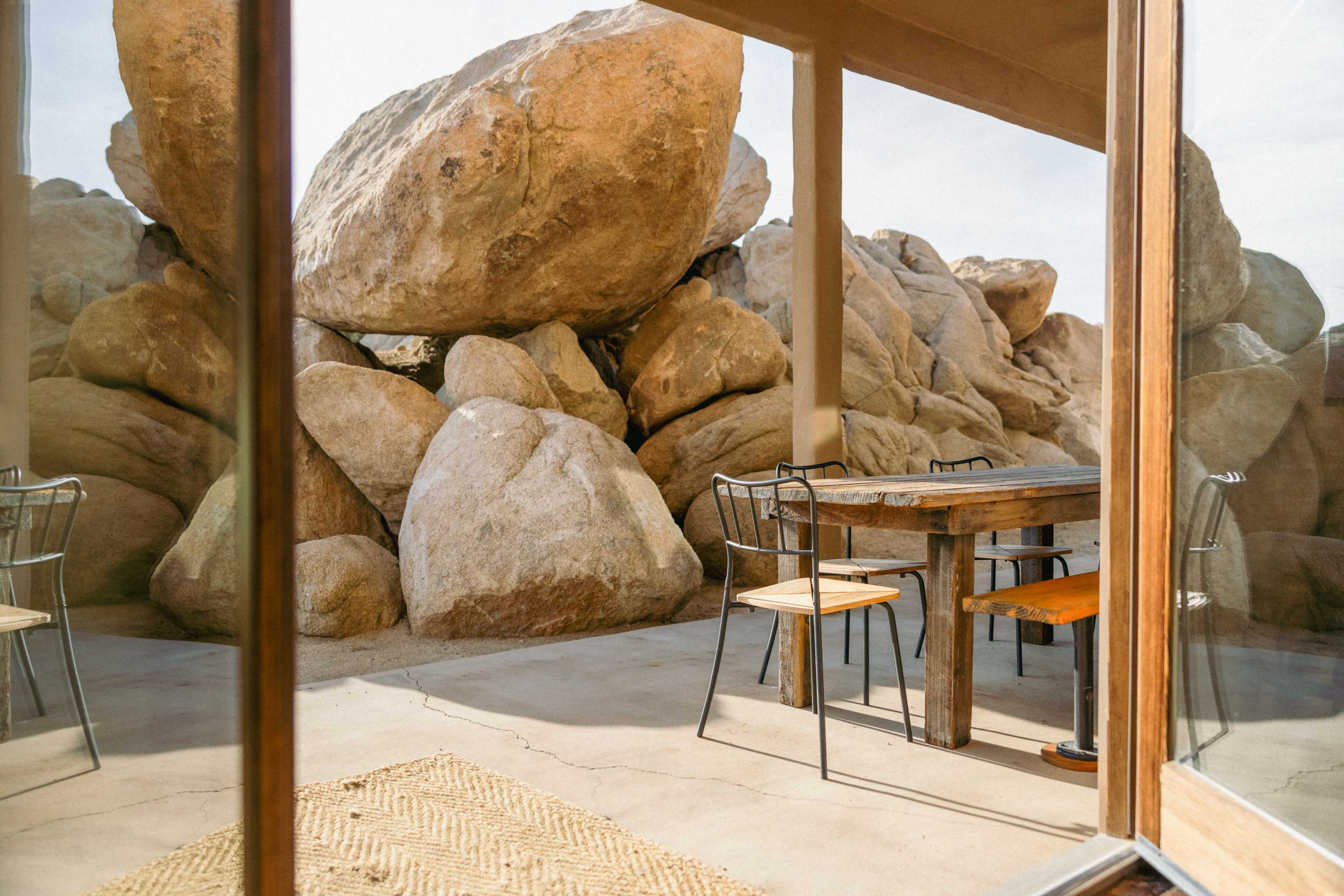 A patio area with a wooden table and chairs is surrounded by large boulders under a clear sky.