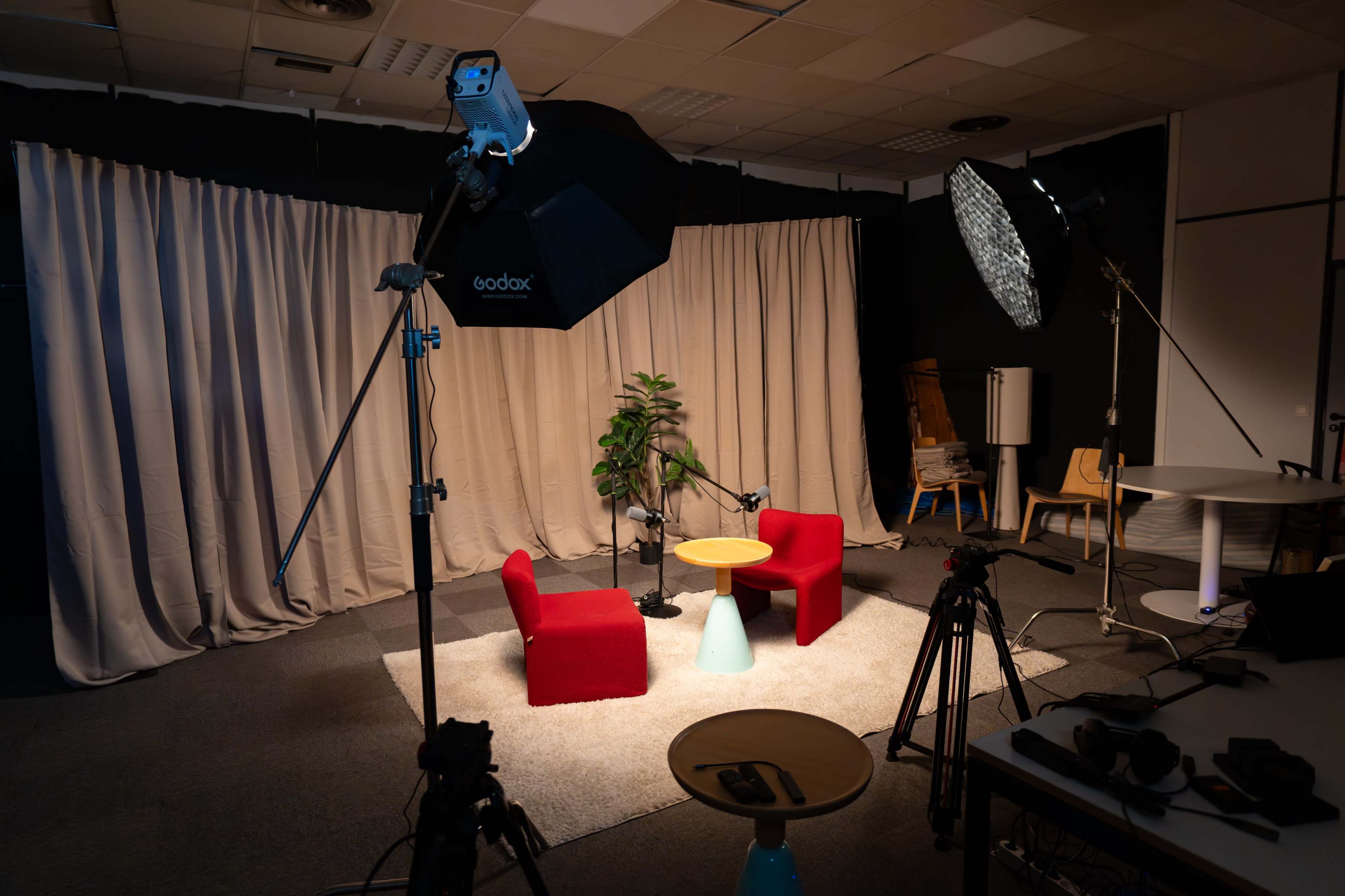 The image shows a dimly lit studio setup featuring two red chairs, a round table, a plant, and multiple lighting equipment in front of a backdrop of curtains.