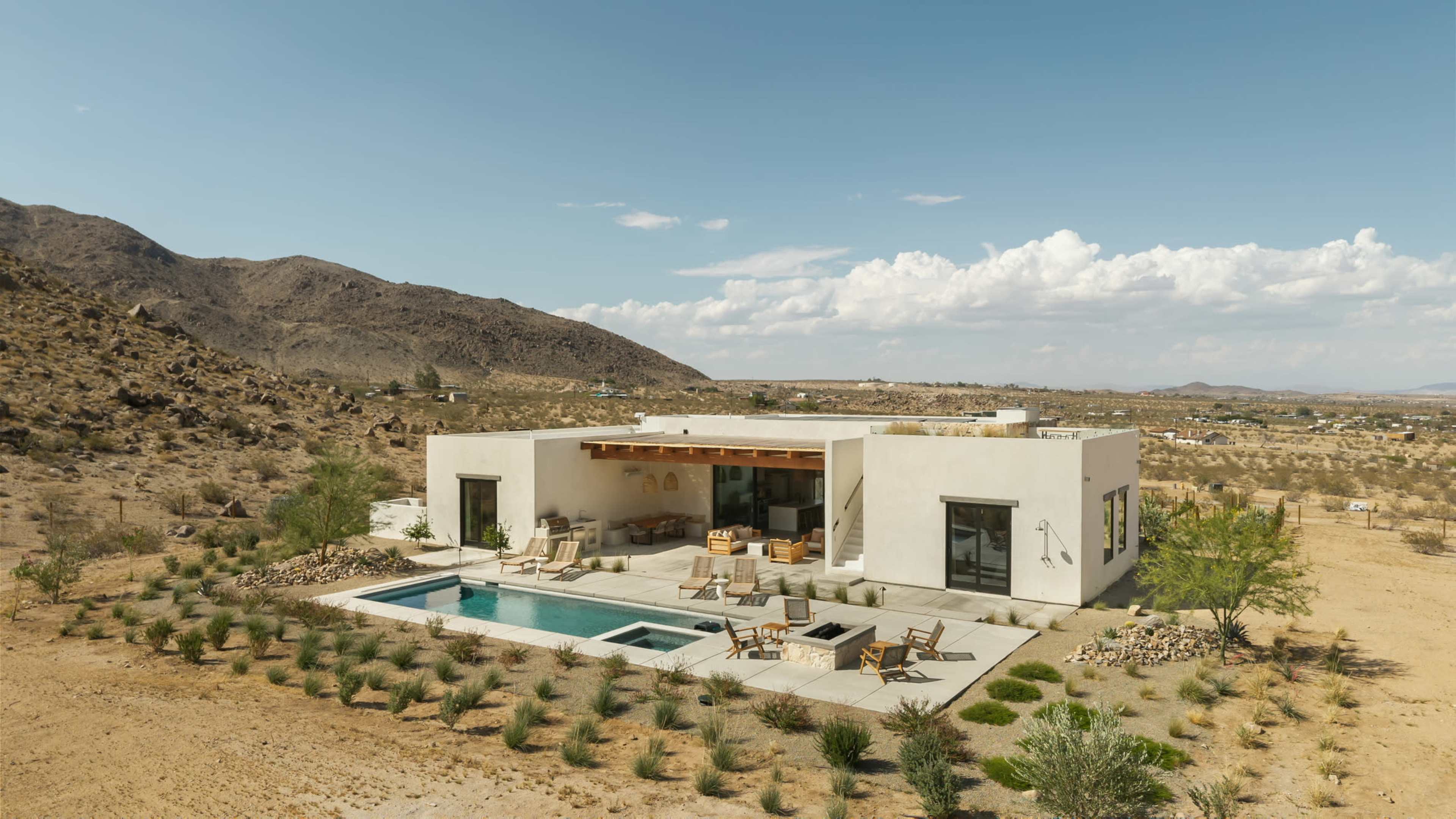A modern, single-story house with a pool is situated in a desert landscape surrounded by mountains and sparse vegetation.