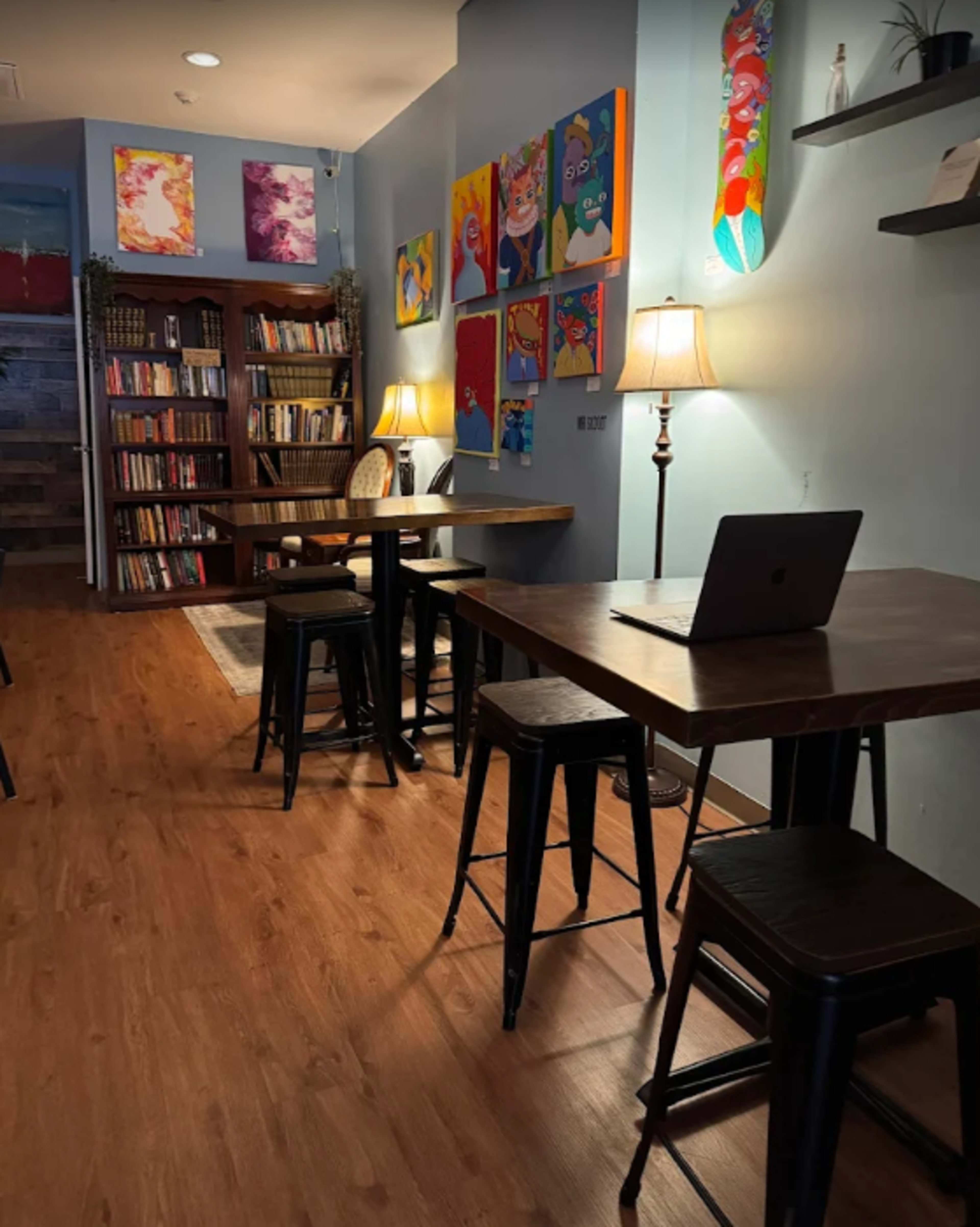 The image shows a cozy café interior with wooden tables, metal stools, bookshelves lined with books, and colorful artwork on the walls, illuminated by lamps.