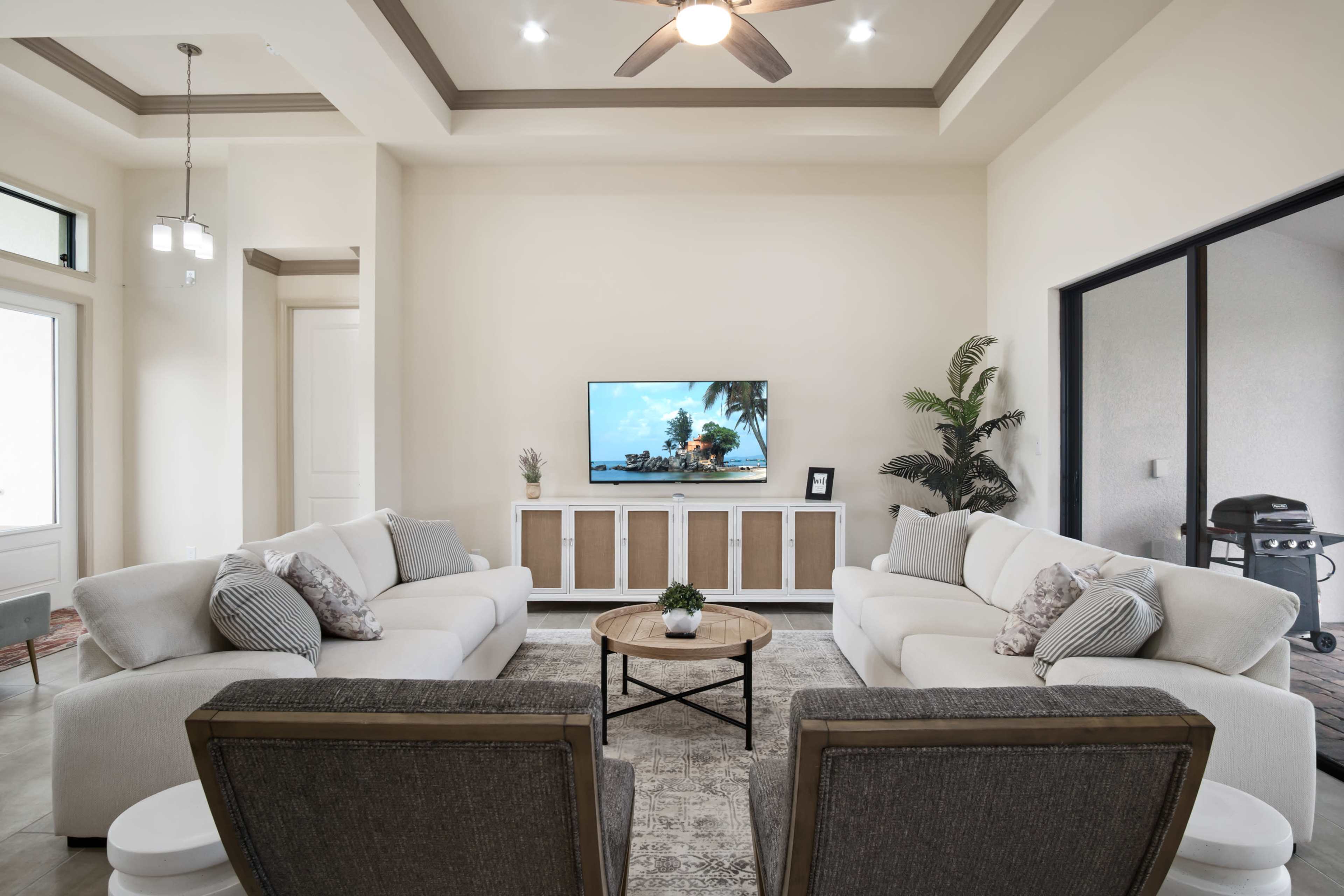 A modern living room features two white sofas and two gray chairs facing a television mounted on the wall, with a round coffee table and a potted plant in the center.