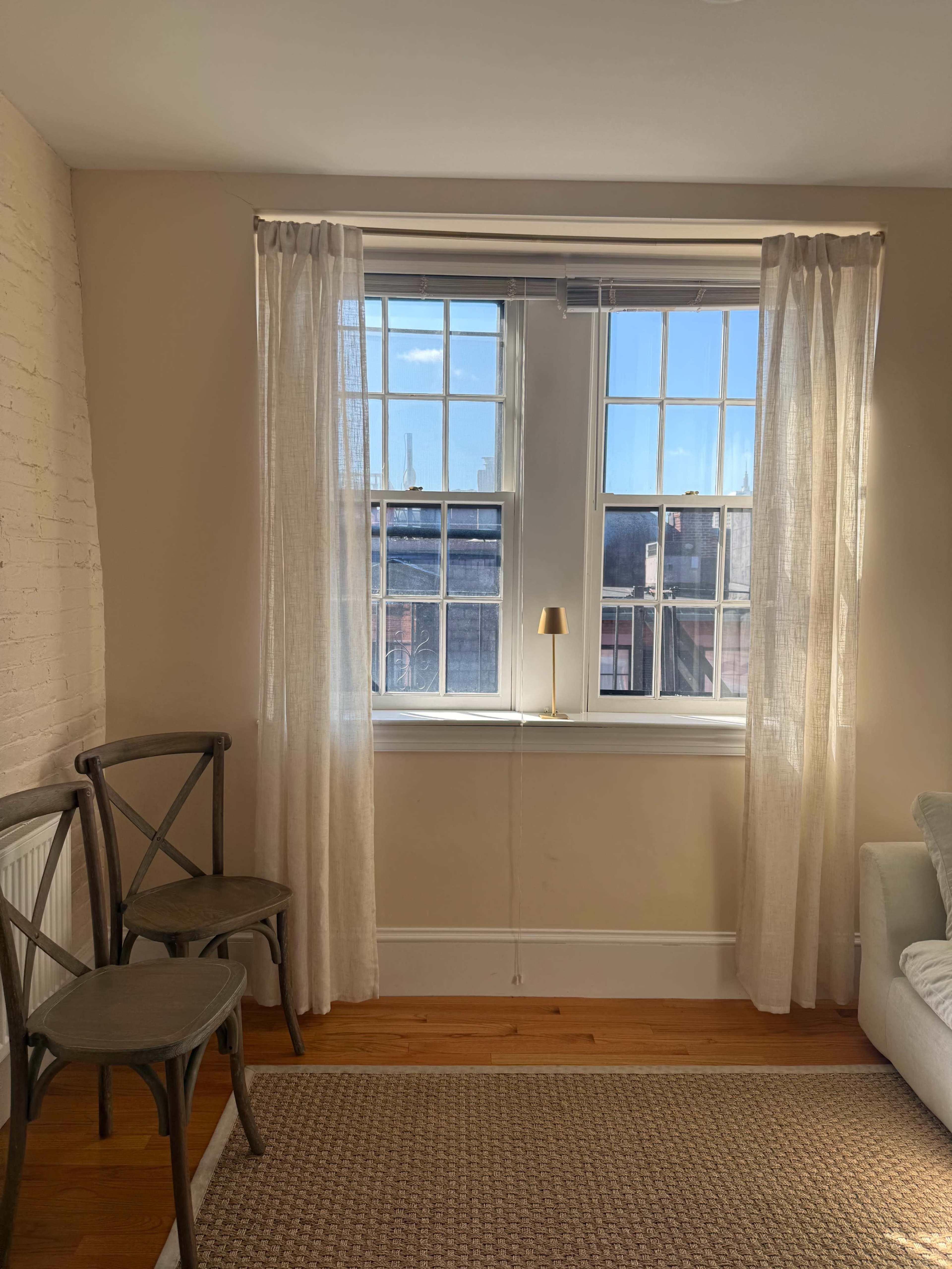 The image shows a bright room with two windows dressed in light curtains, a small lamp on the windowsill, and two wooden chairs beside a beige rug.