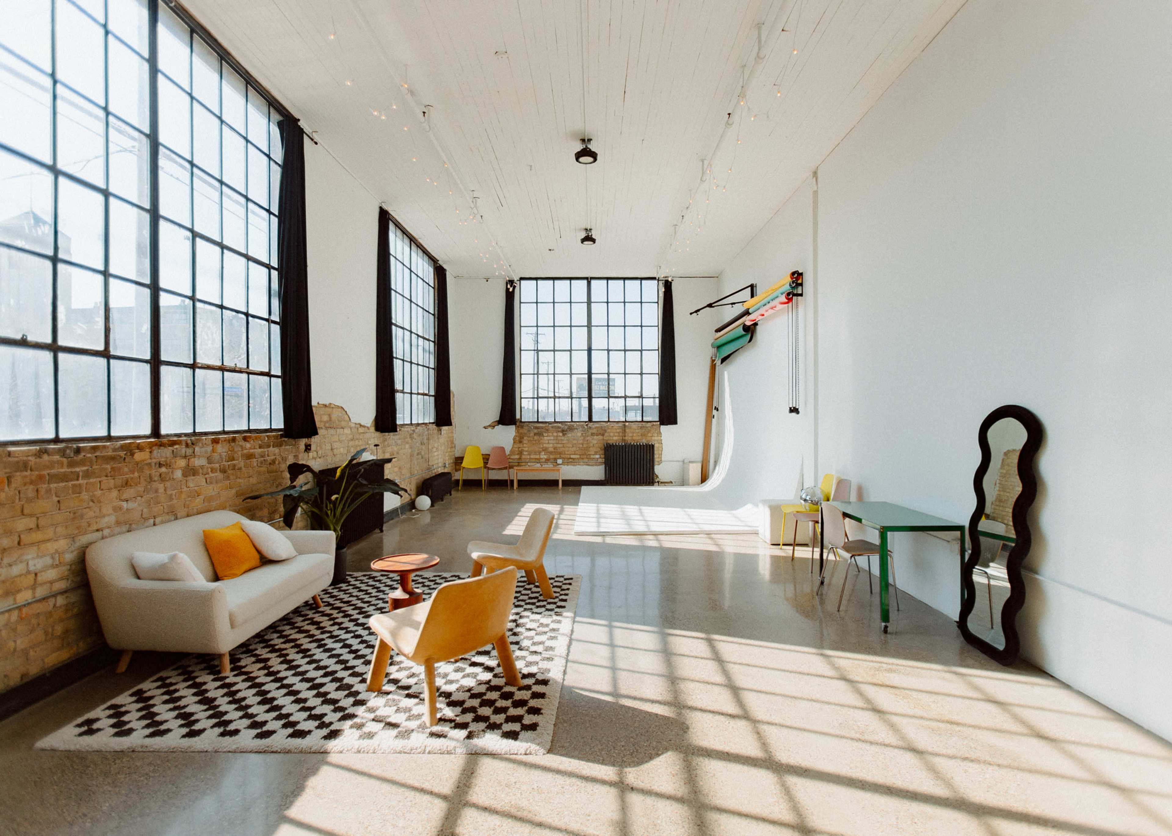 The image shows a spacious, bright studio with large windows, featuring a seating area with modern furniture, a patterned rug, and a backdrop for photography.