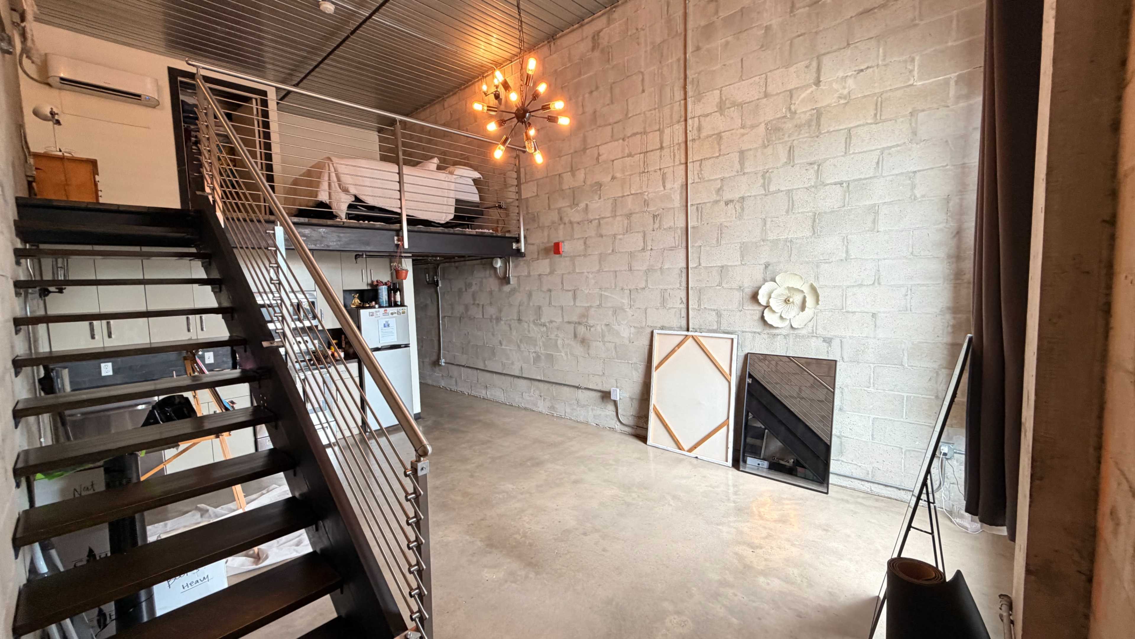 The image shows a spacious industrial-style loft with a staircase leading to an elevated sleeping area, featuring a dining space and a large mirror against the concrete wall.