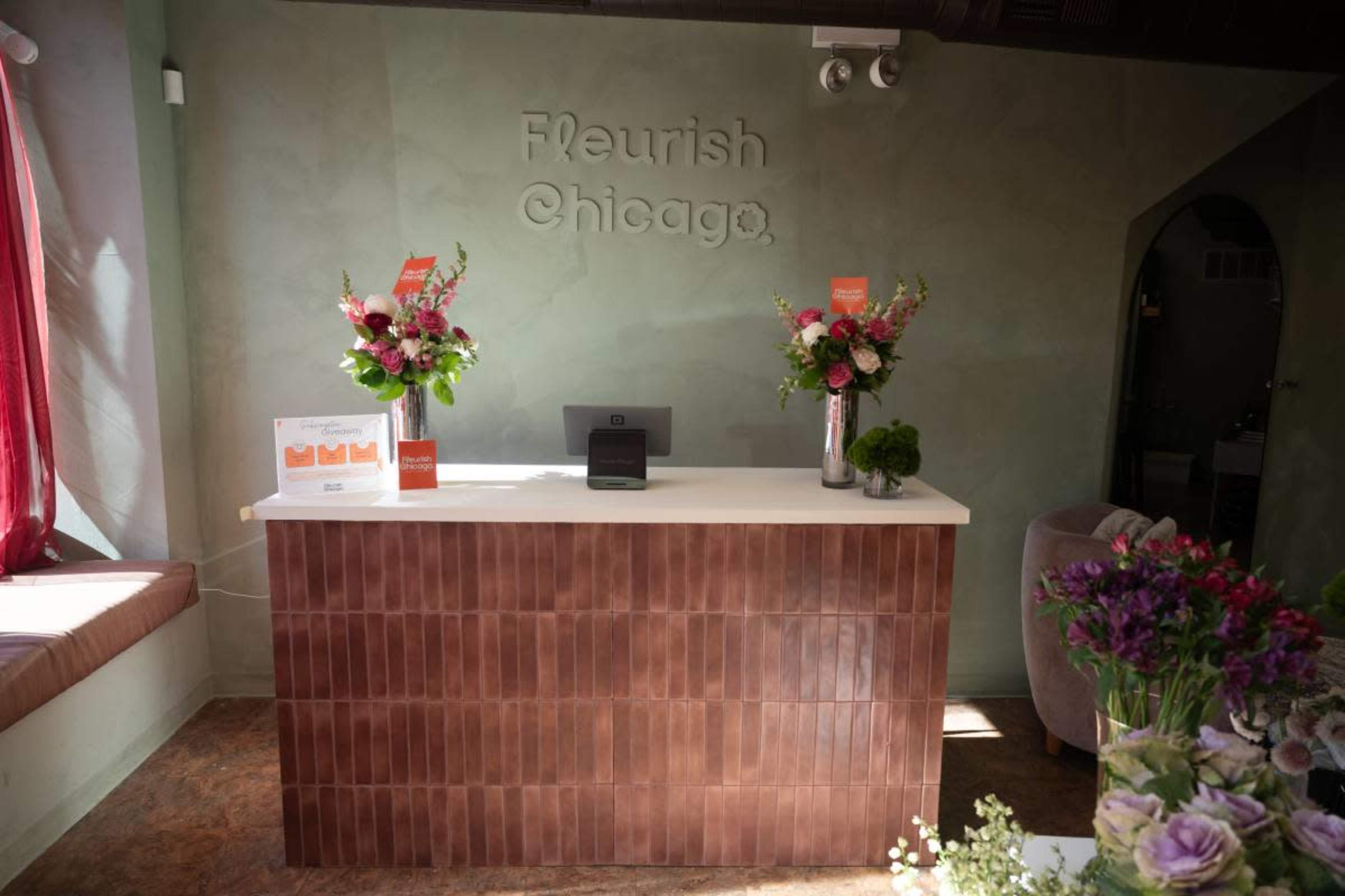 The image shows a reception desk with a textured brown tile front, elegant flower arrangements on either side, and a muted green wall displaying the name "Flourish Chicago."