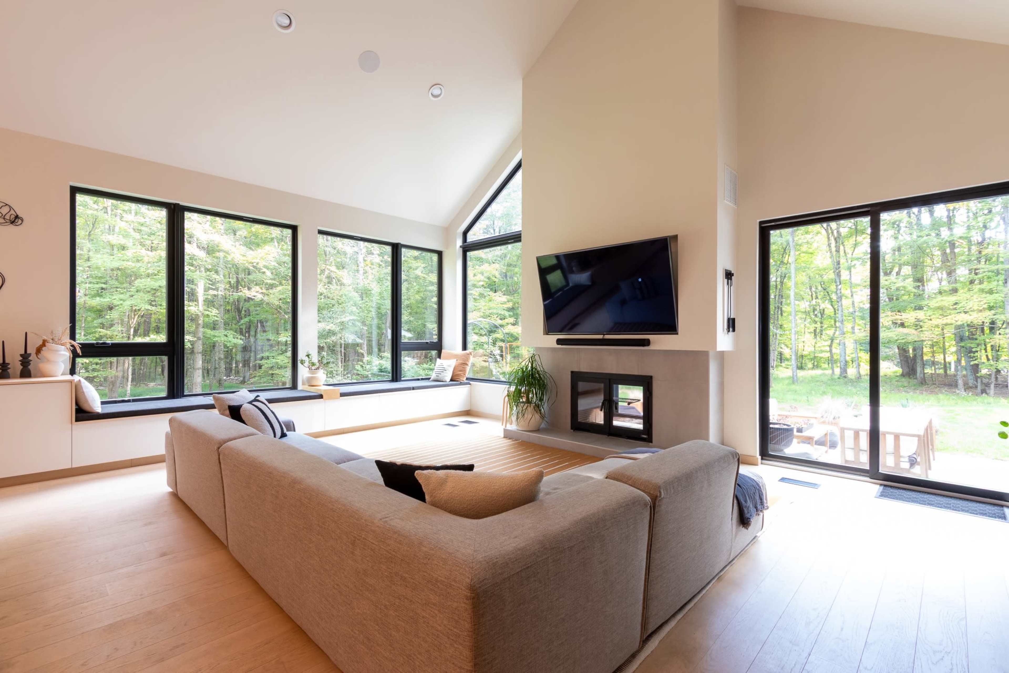 A spacious living room features large windows overlooking a wooded area, with a modern sofa and a mounted television.