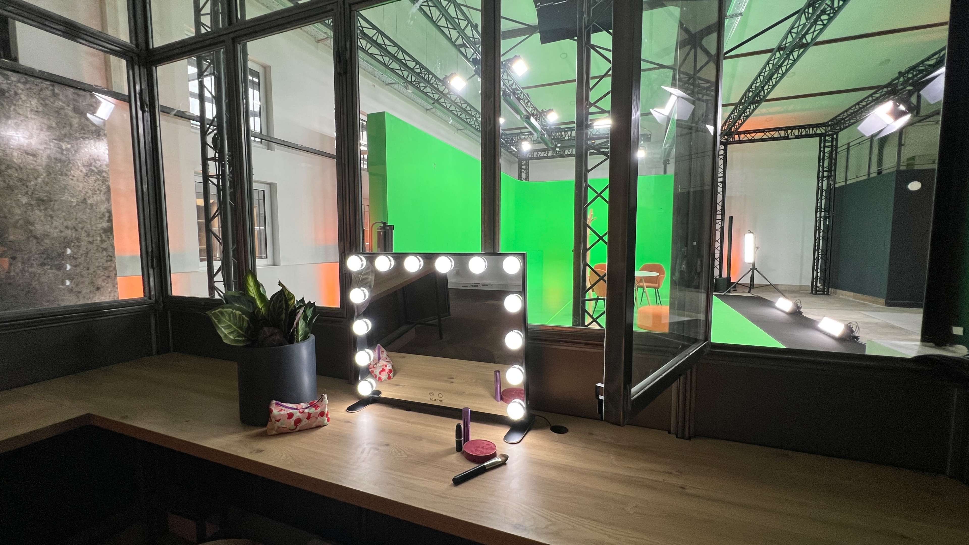 A makeup mirror with lights is placed on a wooden table in front of a glass wall, revealing a green screen studio setup behind it.