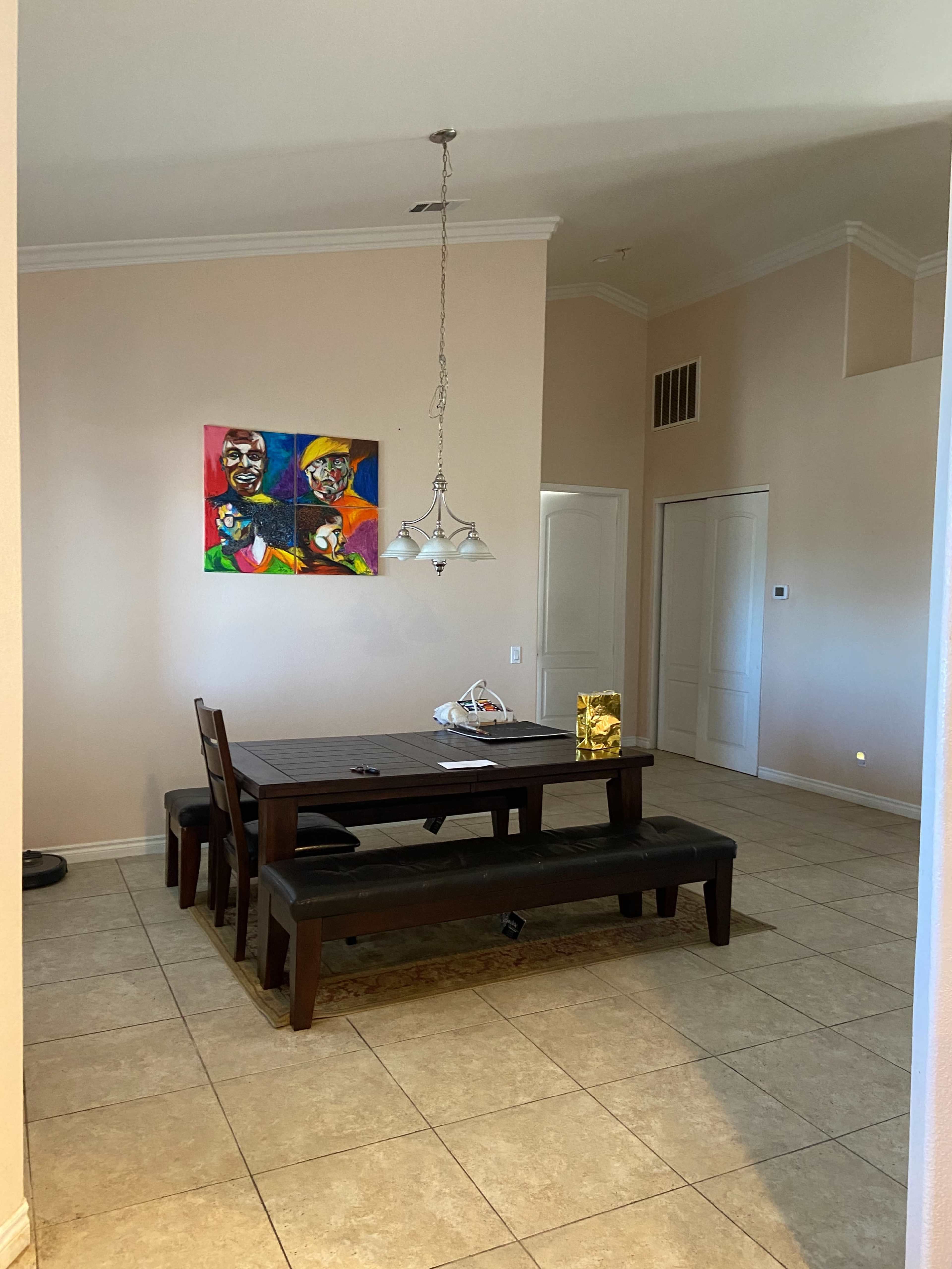 A dining area features a wooden table with benches, surrounded by beige walls and a colorful artwork hanging above.